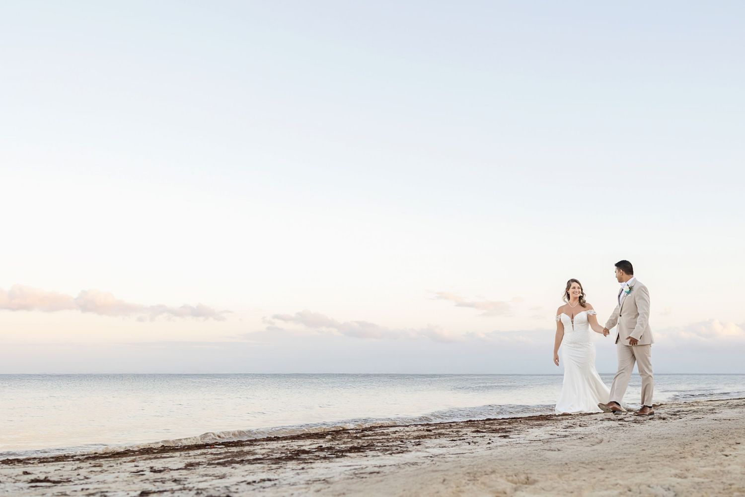 A couple walks hand in hand along a serene beach at sunset wearing elegant white wedding attire.