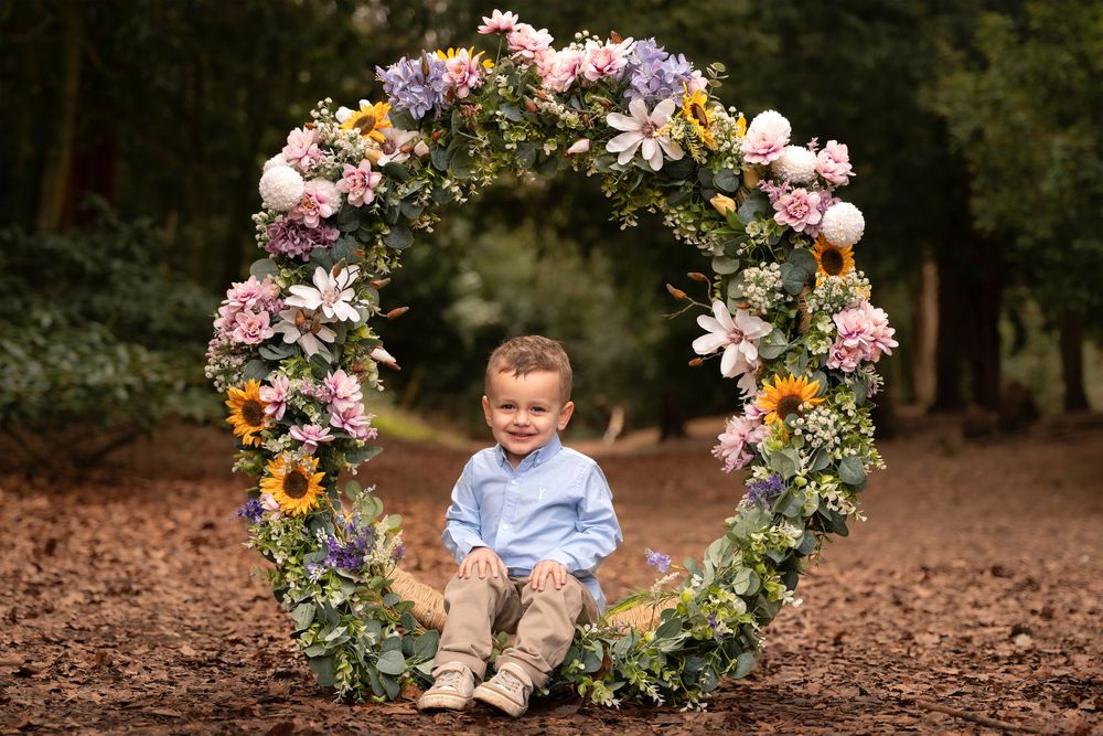 A floral arch with pink and white blooms stands in a wooded outdoor setting during a photo session.