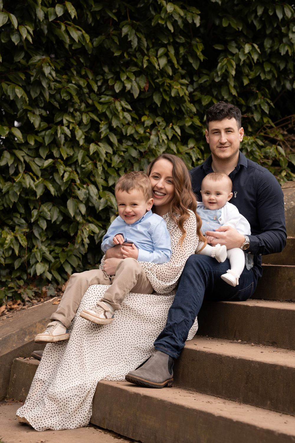 A family sits together on outdoor stone steps with a backdrop of lush green ivy leaves.