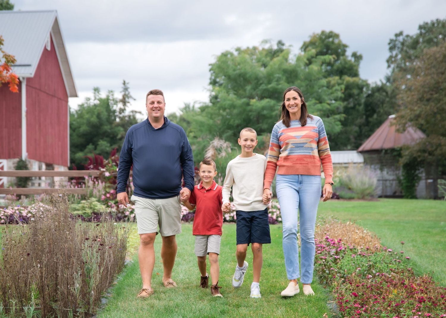Family walking together on a winding garden path lined with colorful wildflowers and red barn in background.