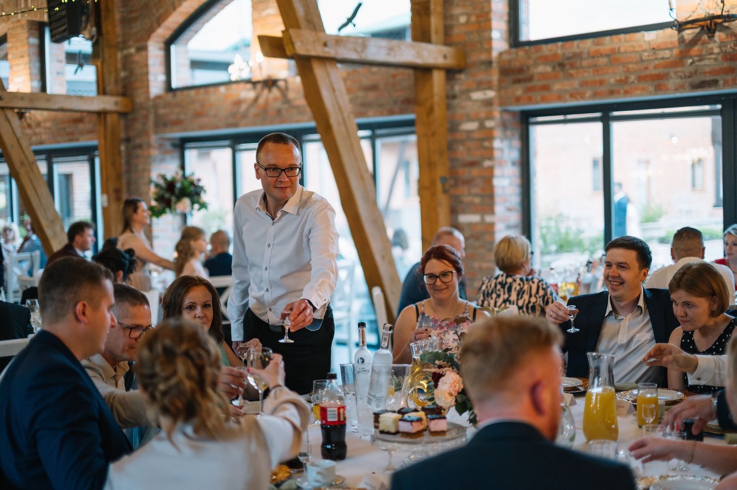 Wedding guests dining in rustic brick reception hall with wooden beam ceiling.