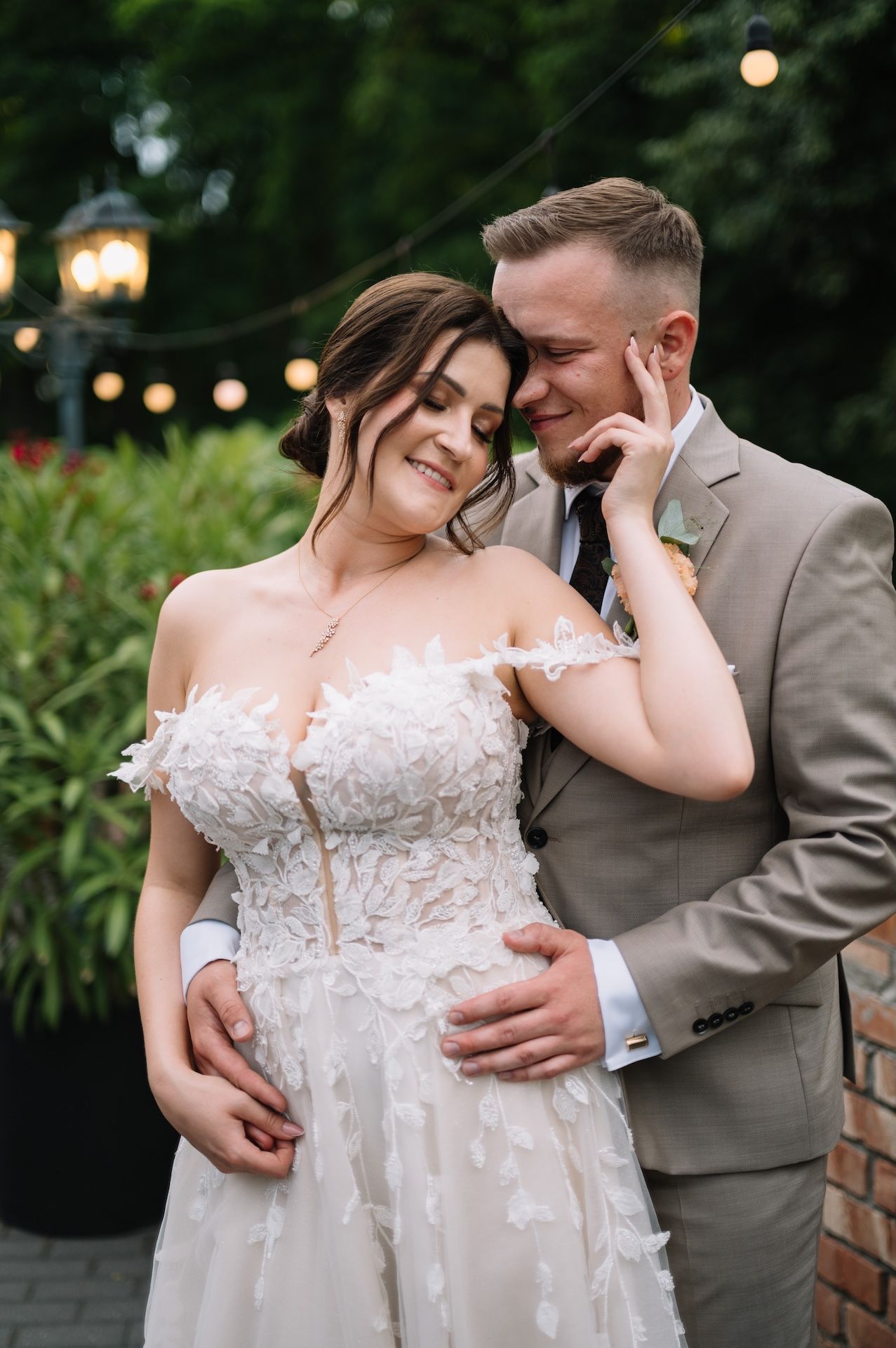 Bride and groom embracing tenderly outdoors under string lights at evening wedding.