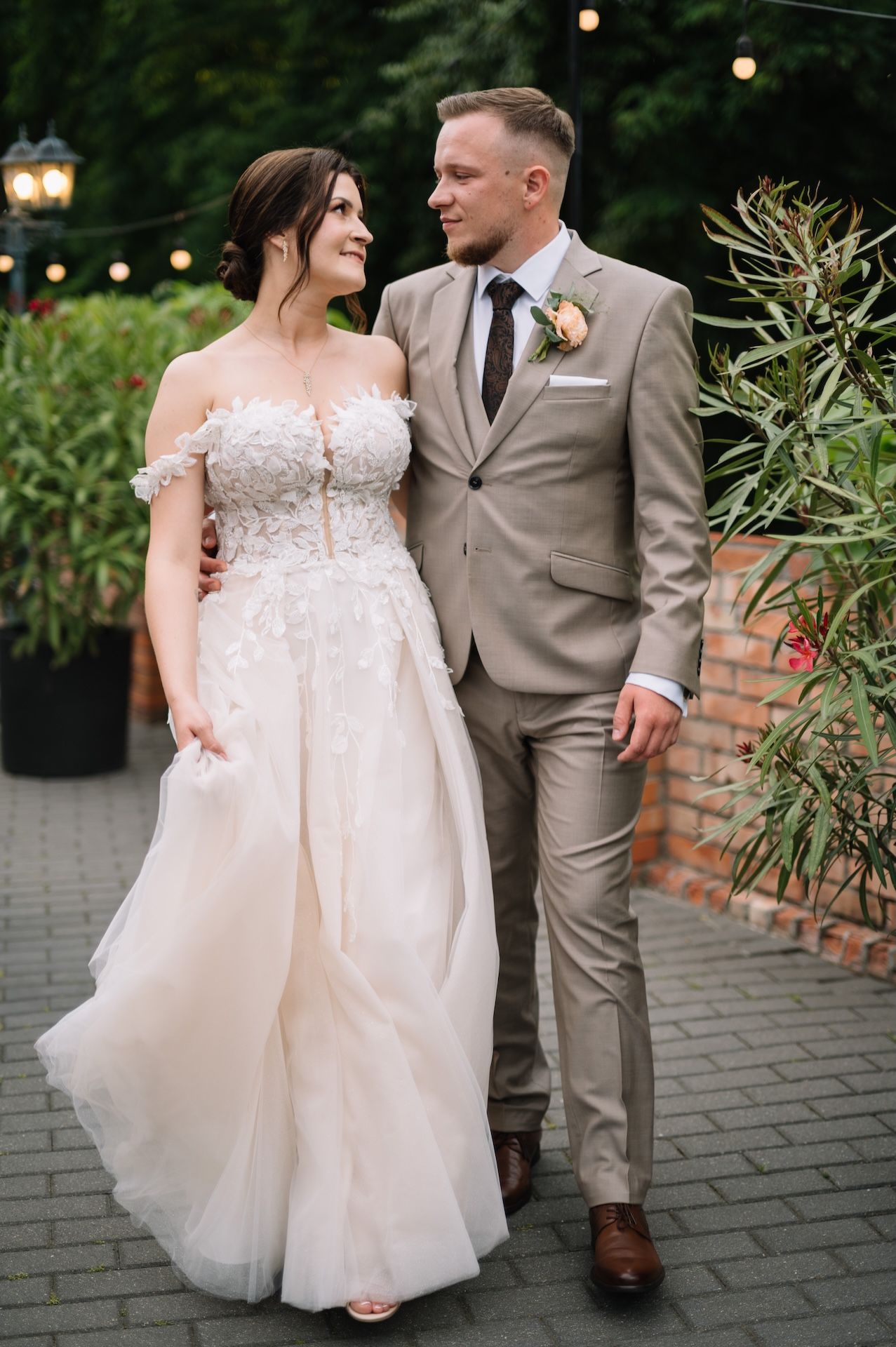 Bride in floral off-shoulder gown and groom in tan suit share a loving glance while walking at outdoor wedding venue.