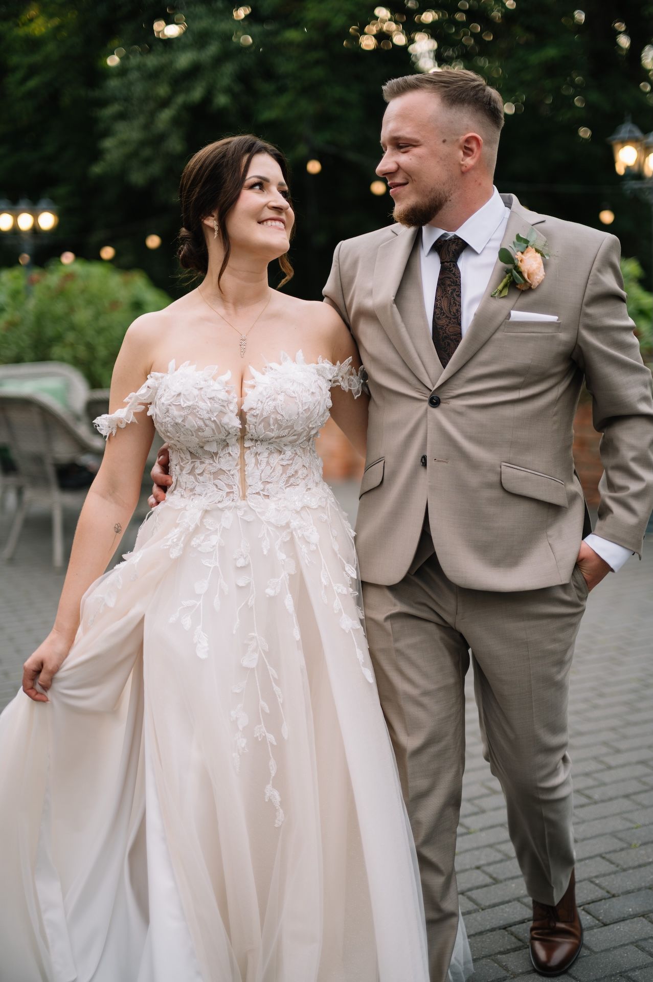 Newlyweds walk together at dusk, bride in white lace off-shoulder dress and groom in beige suit with boutonniere.