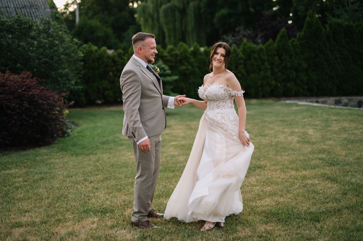Bride and groom hold hands smiling on green lawn, she wears an elegant off-shoulder floral wedding gown.