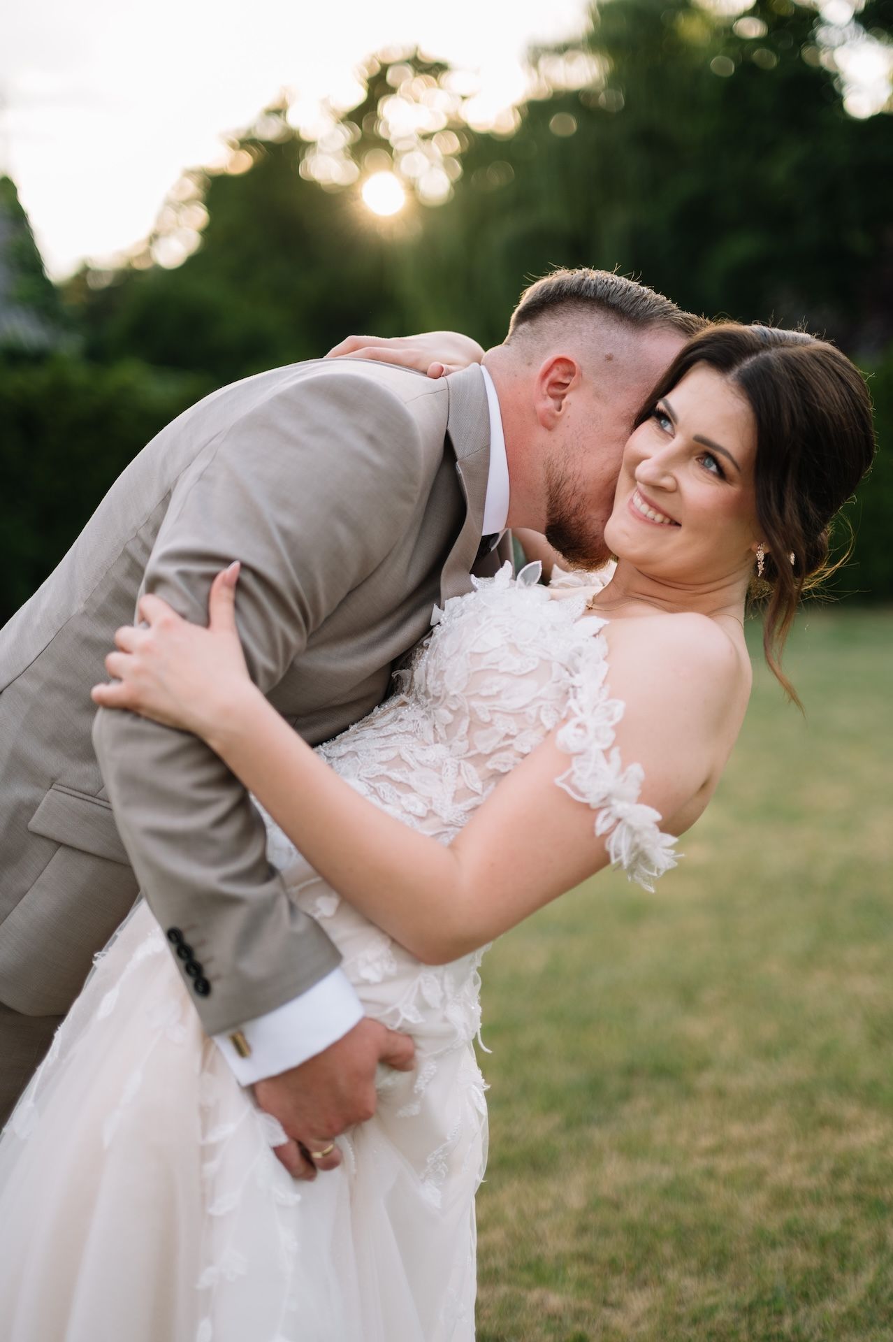 Groom in gray suit kisses bride on cheek as she smiles joyfully, embracing during golden hour outdoor wedding.