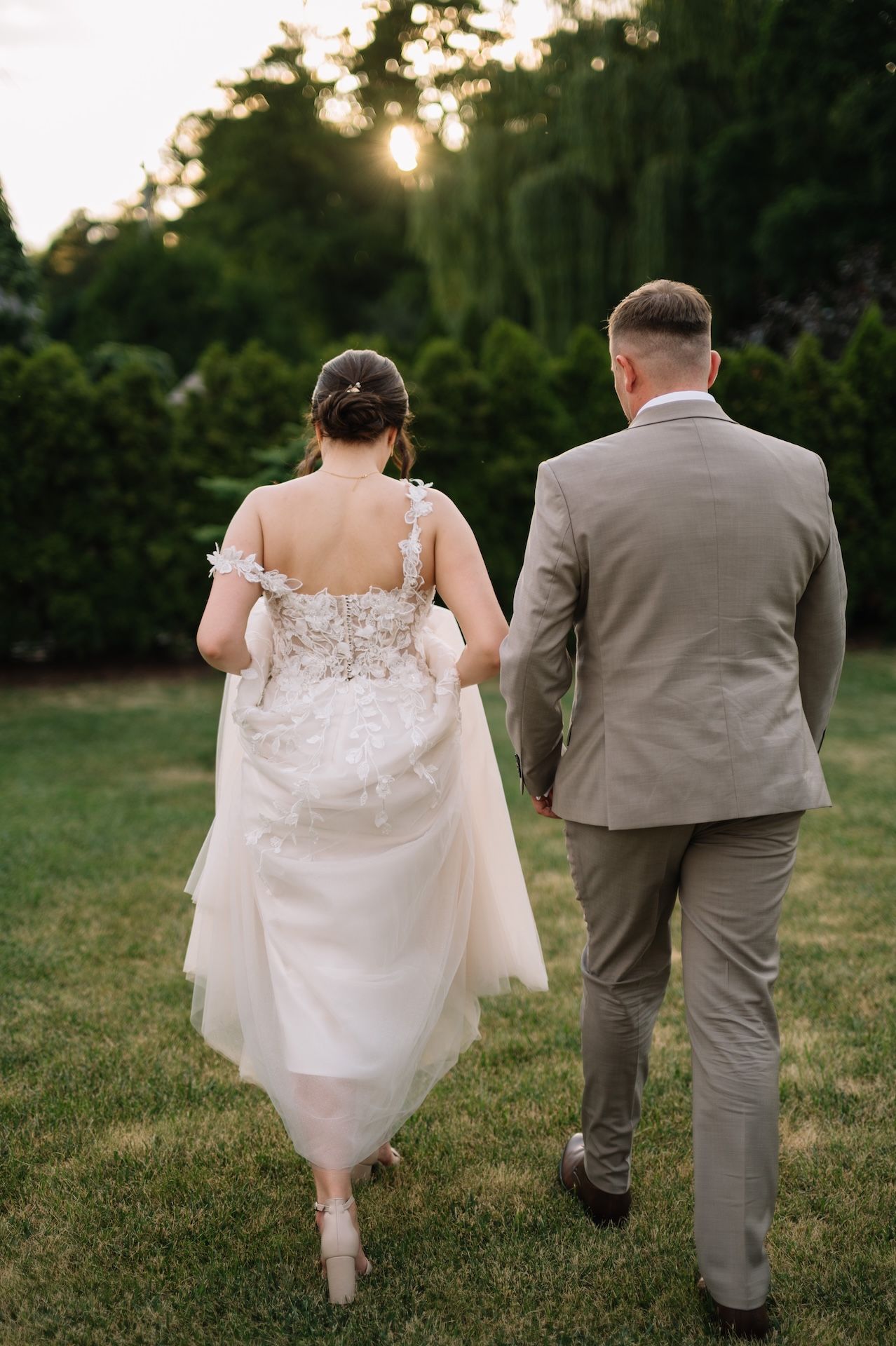 Bride and groom walk hand in hand away from camera at sunset through lush green garden.