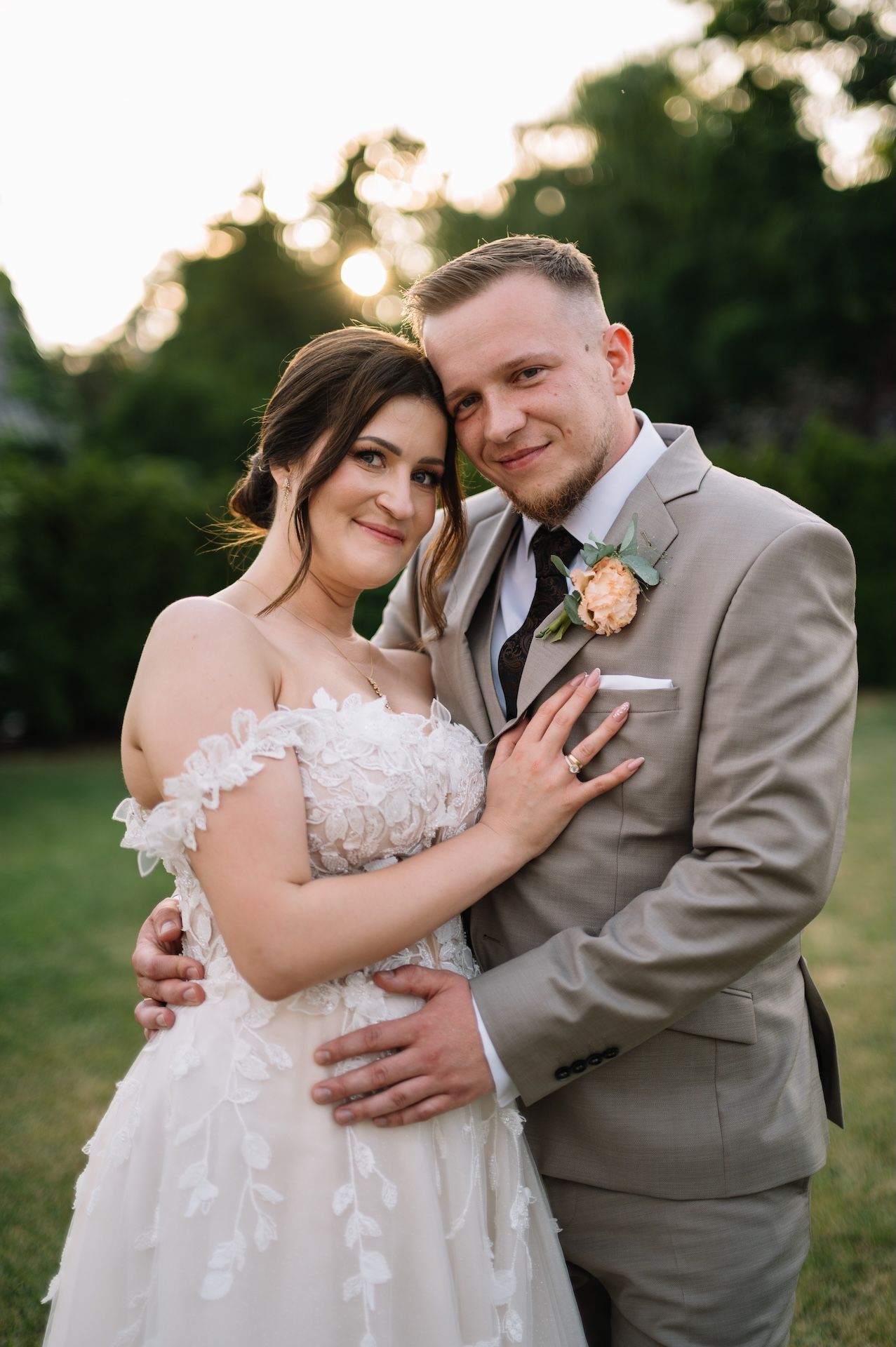 Bride and groom embrace tenderly in warm sunset light, both smiling softly for romantic wedding portrait.