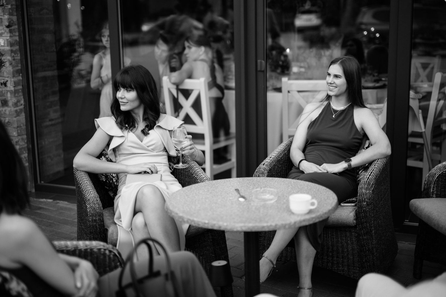 Two elegantly dressed women sit at an outdoor cafe table in black and white wedding reception photo.