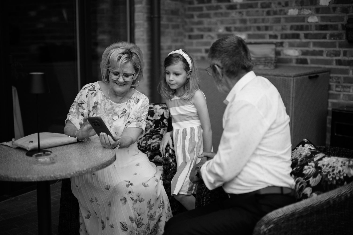 Black and white photo of woman and child looking at phone with older man at wedding reception.