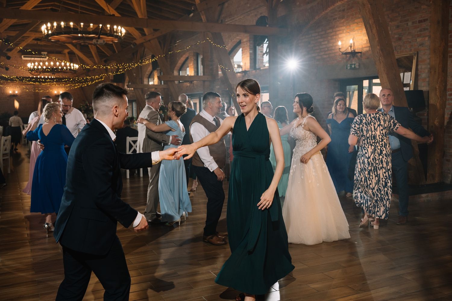 Wedding guests dance joyfully in rustic wood-beamed reception hall with warm string lights overhead.