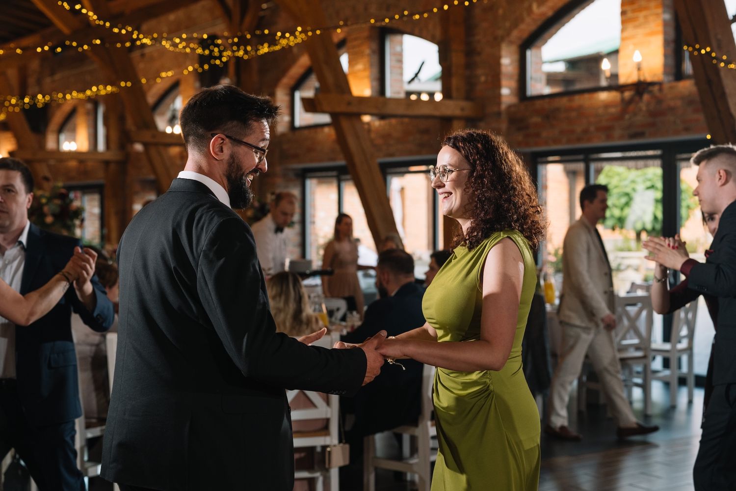 Man in suit and woman in green dress share a handshake and smile at an elegant wedding reception.