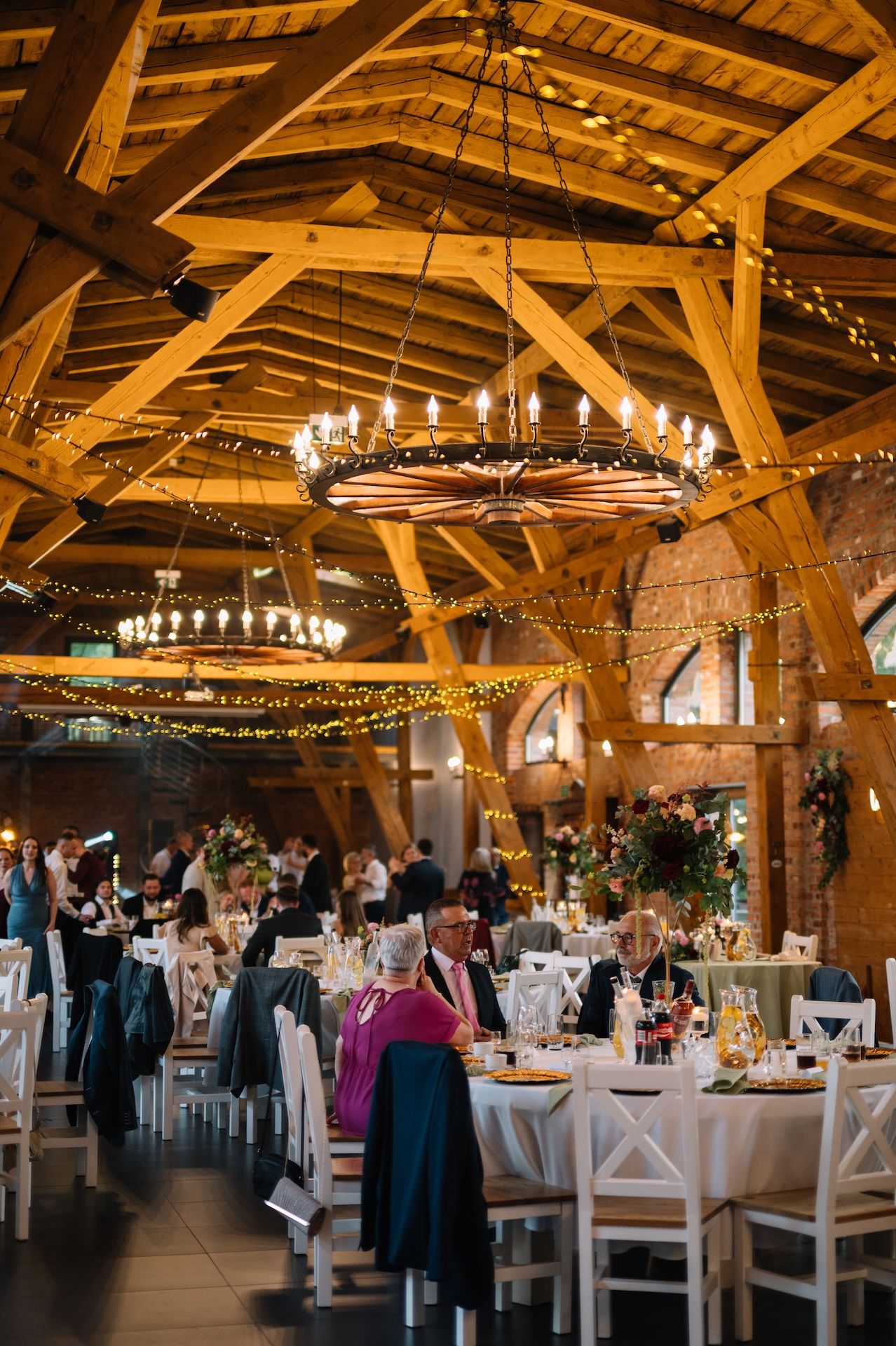 Elegant barn wedding reception with wooden beam ceiling, chandelier, fairy lights, and guests at round tables.
