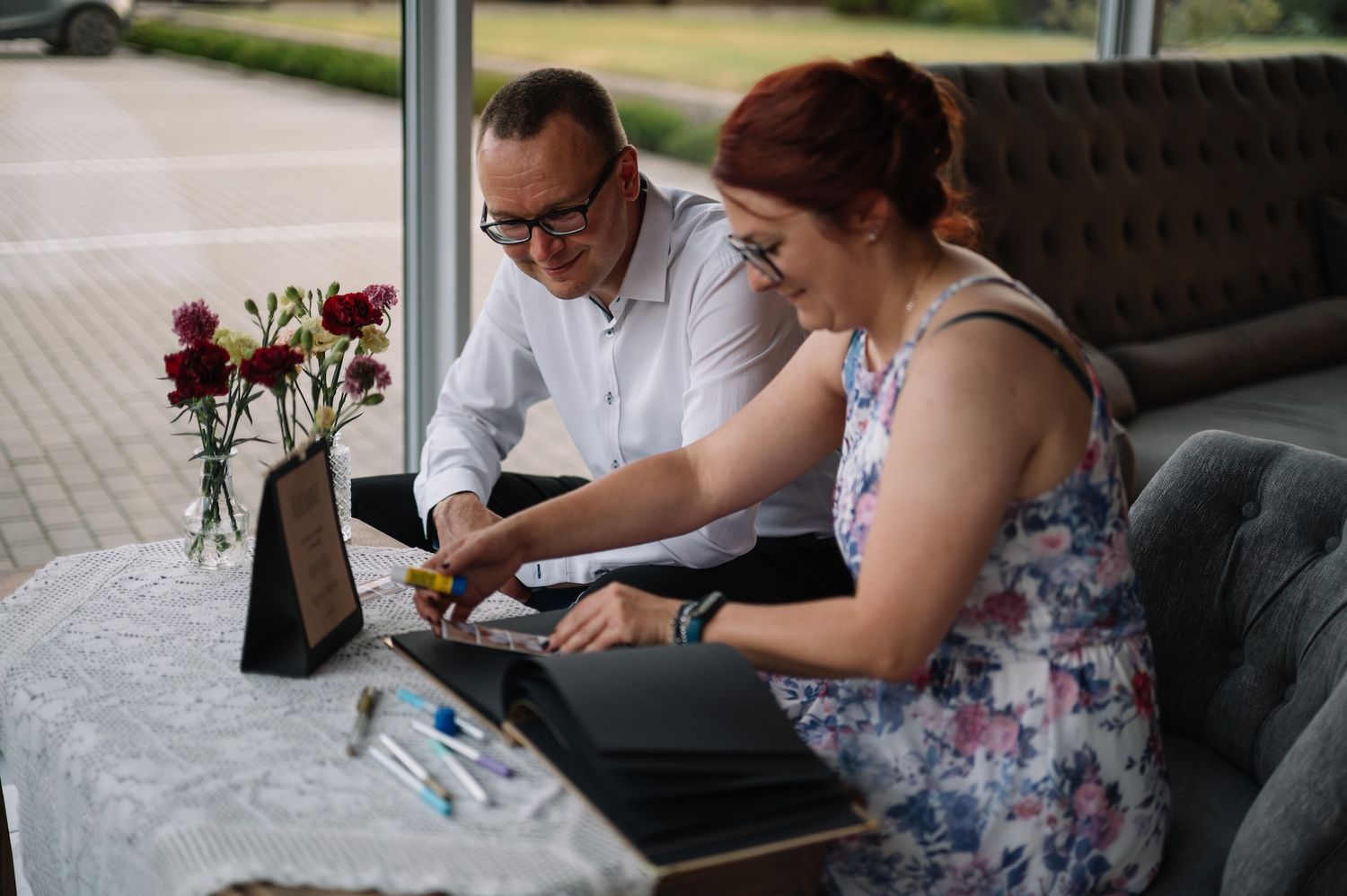 Man and woman collaborating on a laptop at a small table decorated with red roses near large windows.