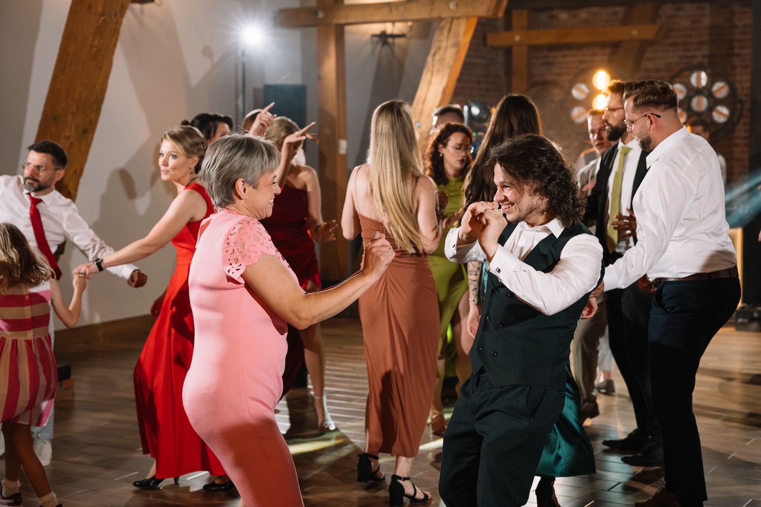 Wedding guests of all ages dancing together joyfully in a rustic brick-walled reception venue.