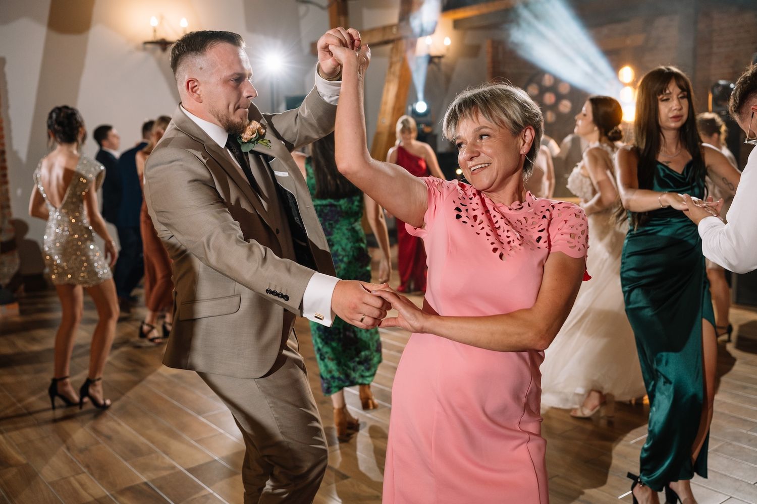 Groom in tan suit dancing with woman in pink dress at wedding reception.