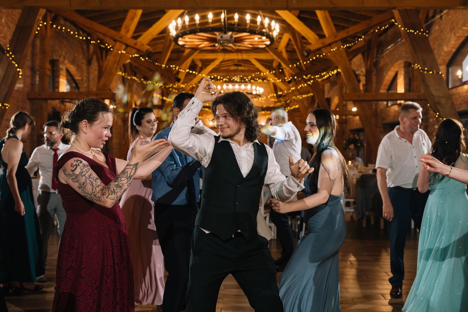 Man in black vest dancing energetically with guests at rustic barn wedding reception.