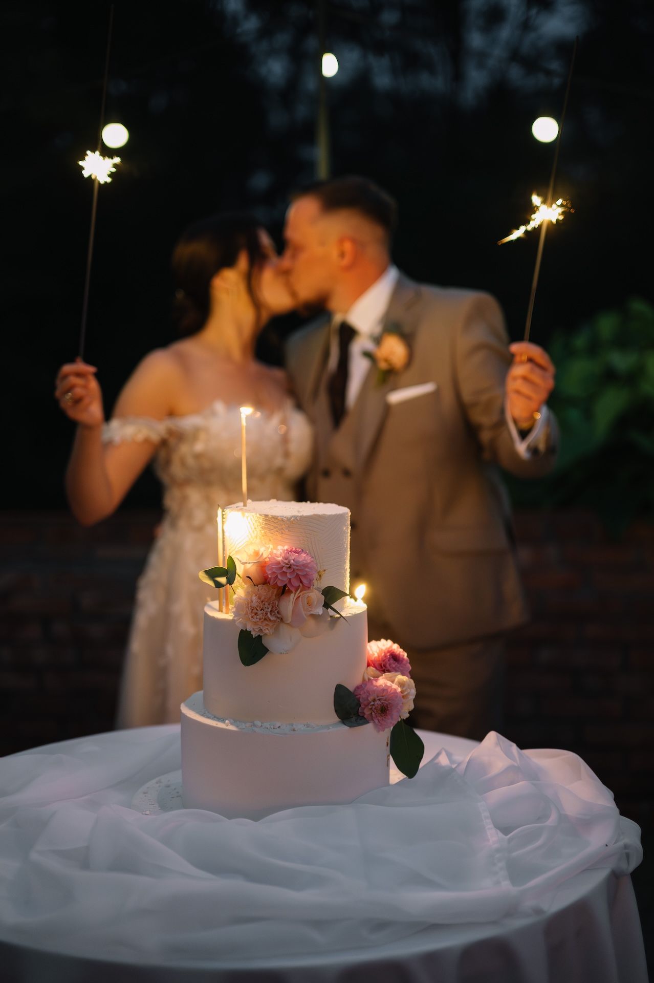 Newlyweds kissing behind three-tier white wedding cake with sparklers at night.