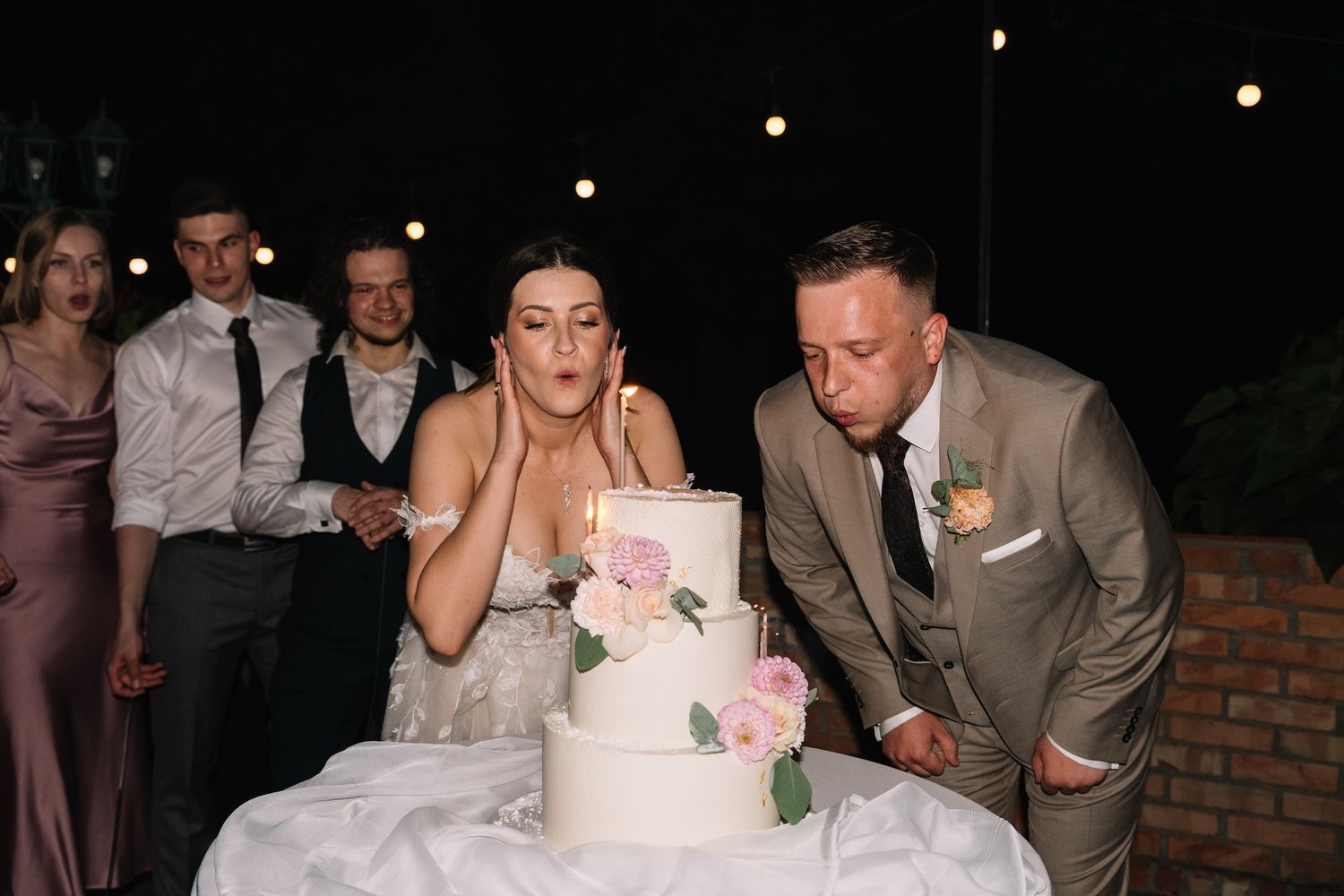 Bride and groom blowing out candles on white three-tier wedding cake outdoors.