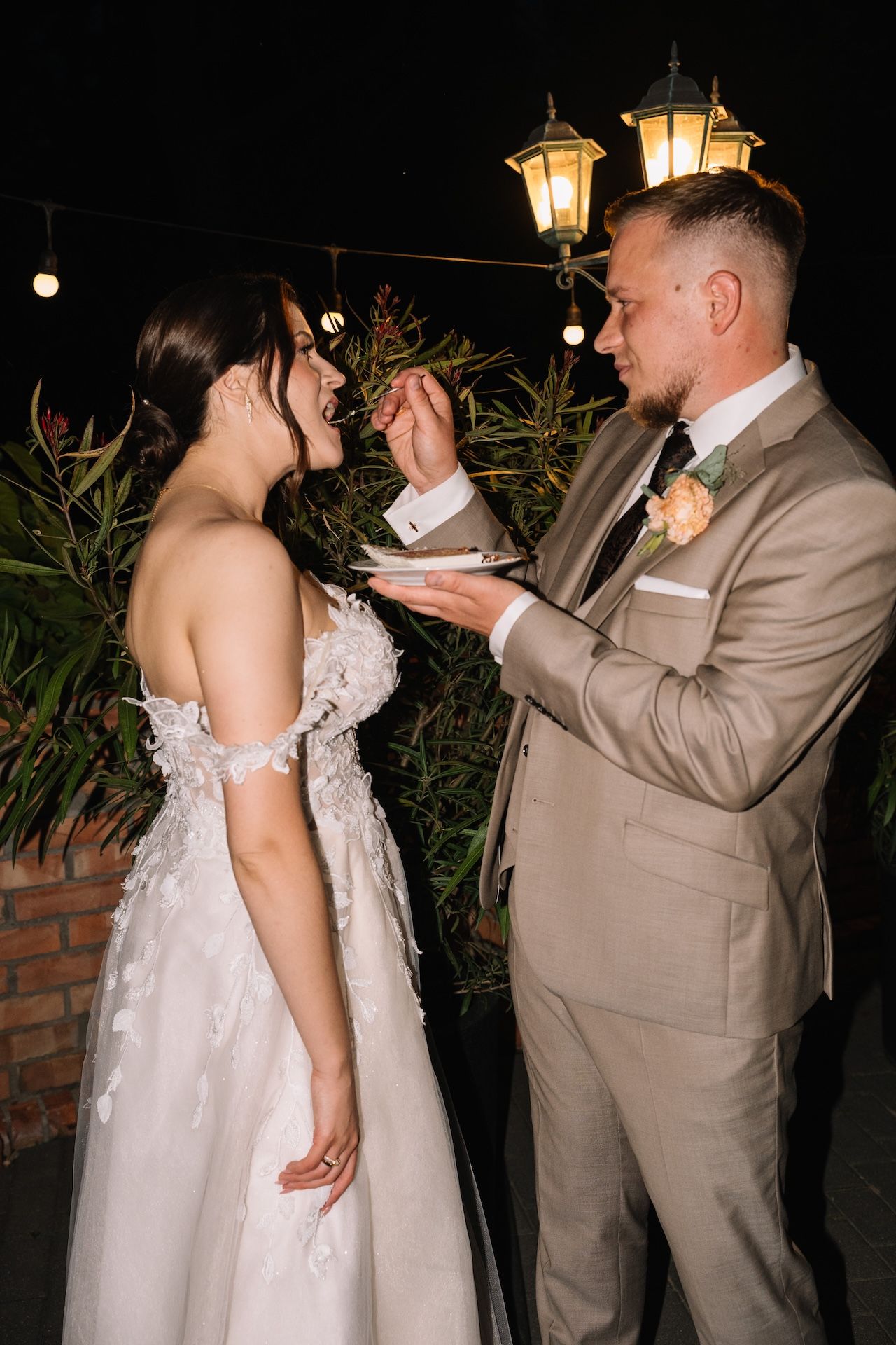 Bride and groom share a sweet cake-feeding moment outdoors at night under warm glowing lanterns.