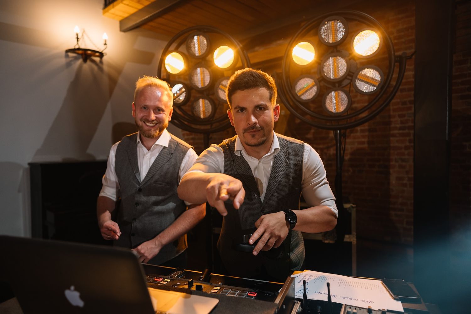 Two smiling DJs operate equipment and laptop at a lively wedding reception venue.