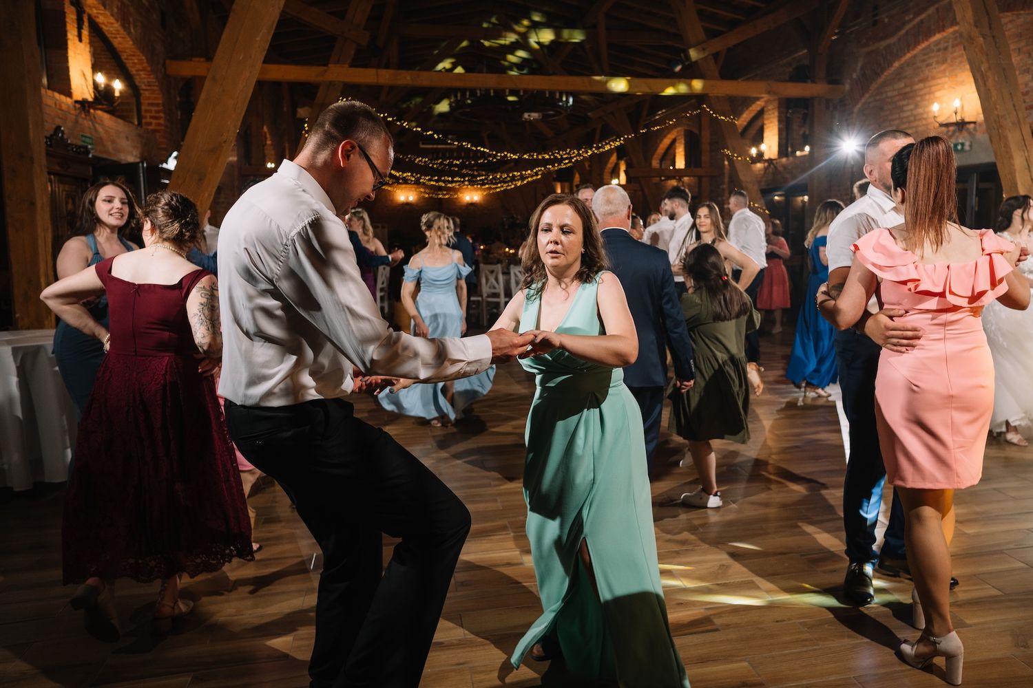 Energetic wedding guests dance together in a rustic barn reception hall with string lights.