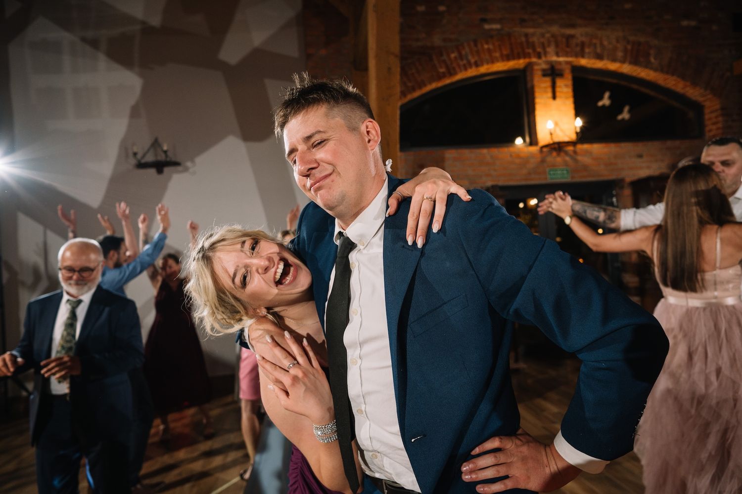 Joyful woman hugs smiling man in blue suit on a lively wedding reception dance floor.