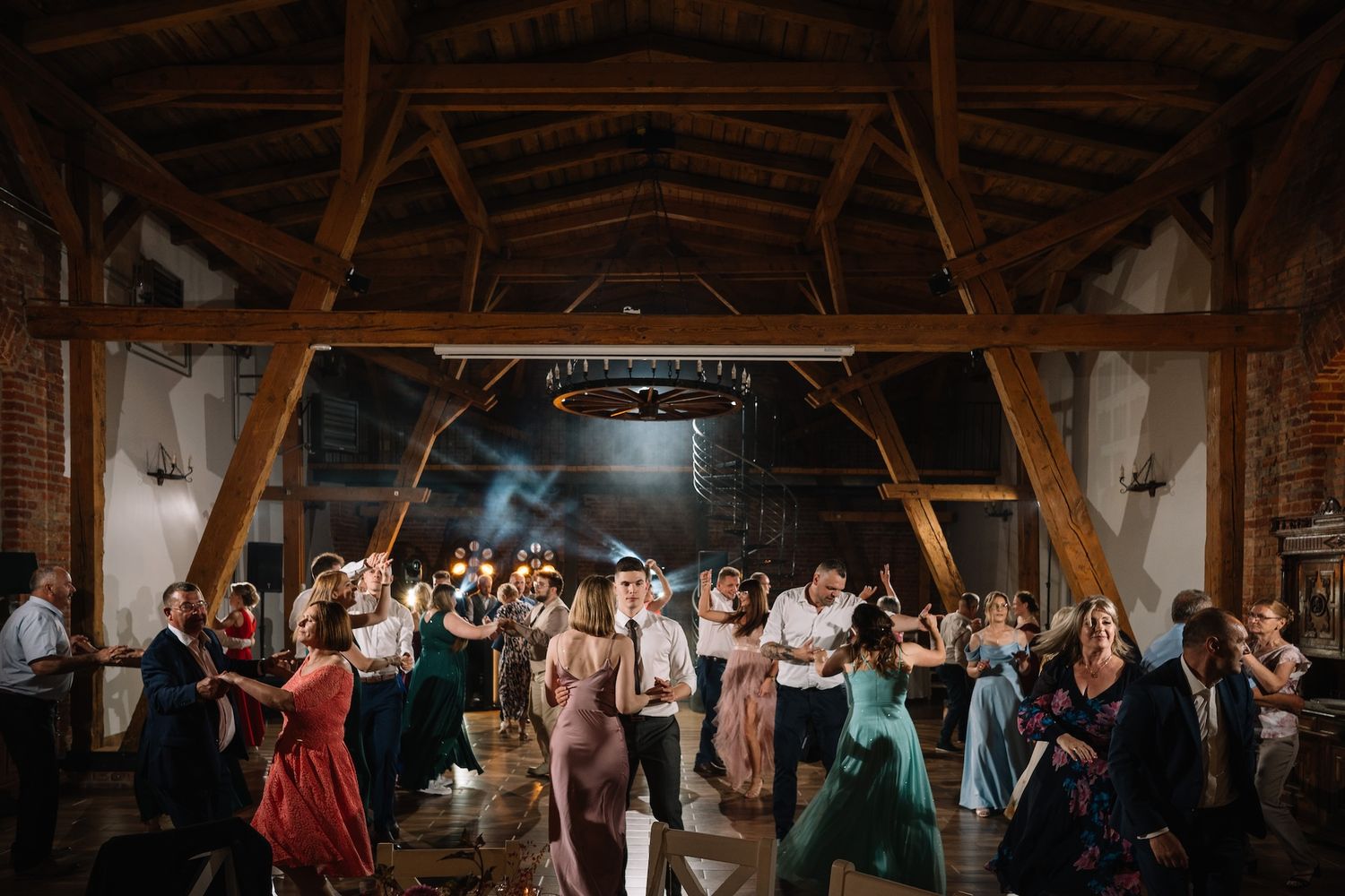 Wide shot of wedding guests filling a rustic barn dance floor under wooden beams and lights.