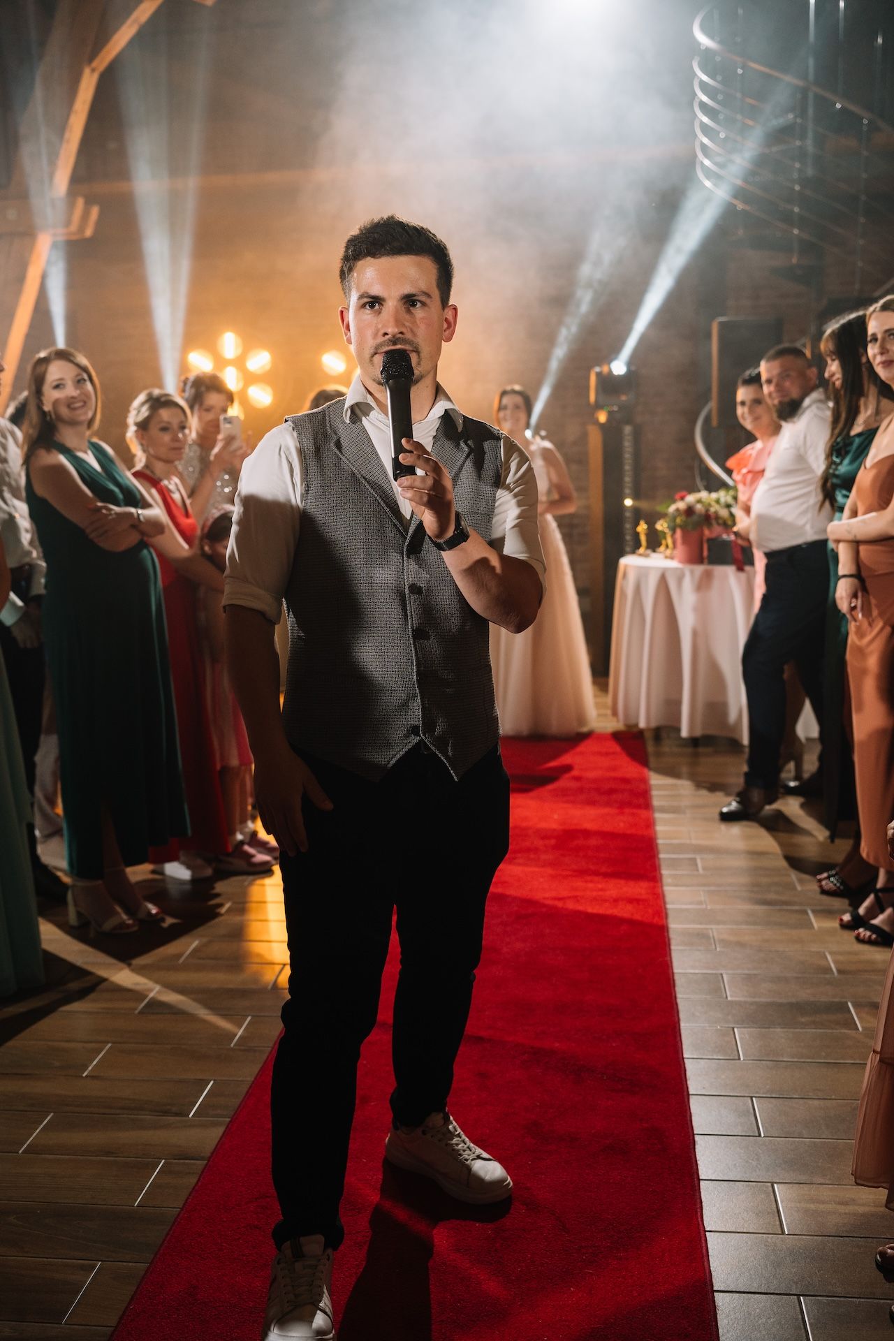 Wedding MC in vest holds microphone on red carpet in a dramatic spotlight-lit reception hall.
