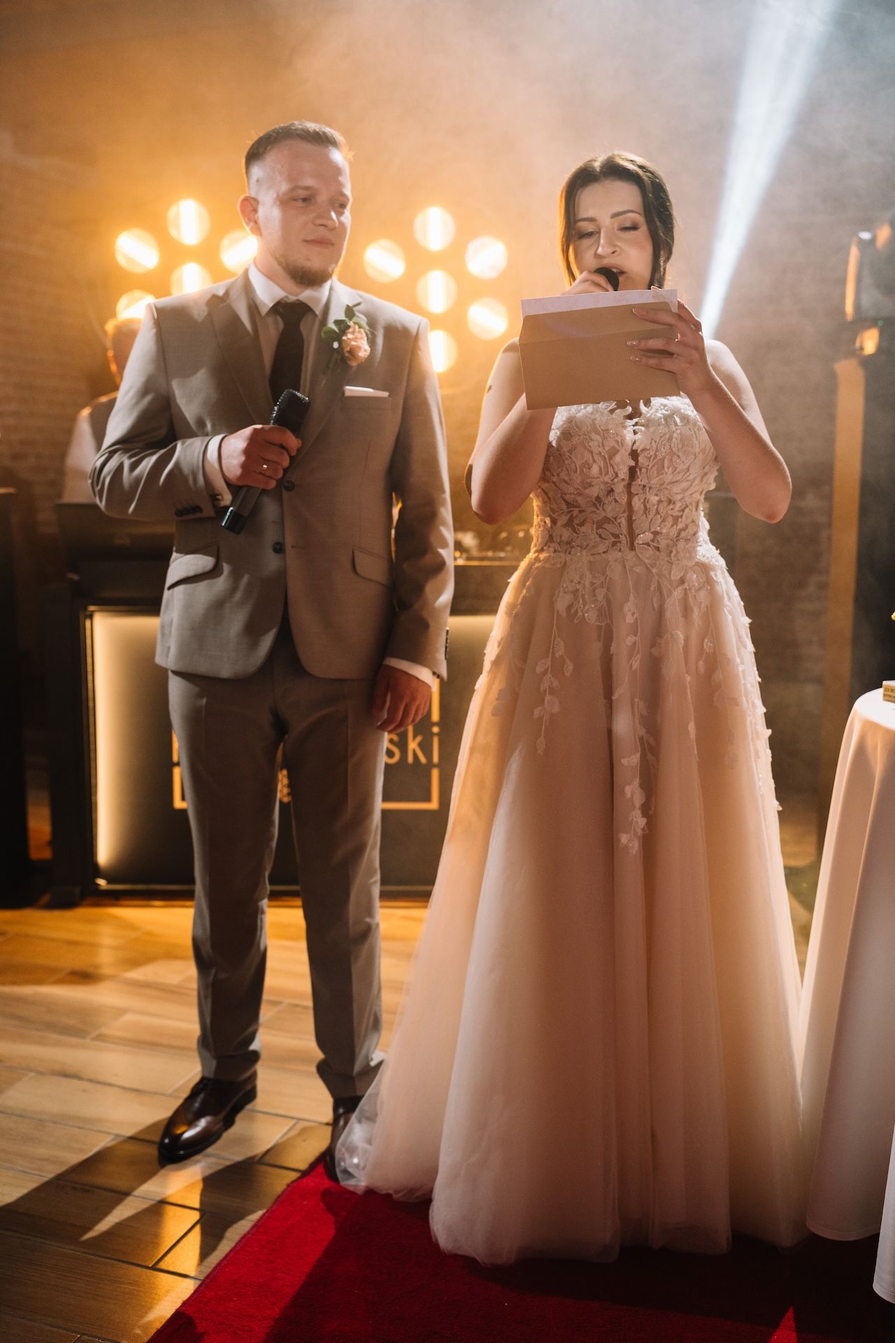 Bride reads vows from paper while groom holds microphone on red carpet at wedding reception.