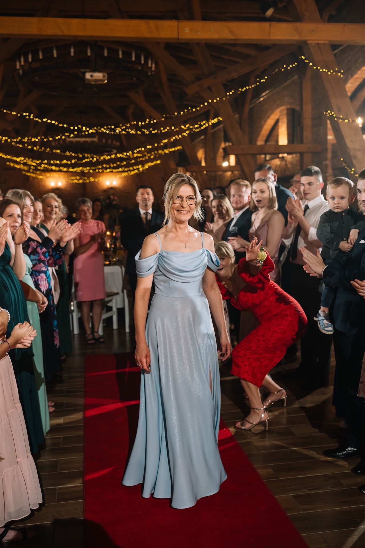 Woman in light blue gown walks red carpet aisle at elegant barn wedding reception with fairy lights.