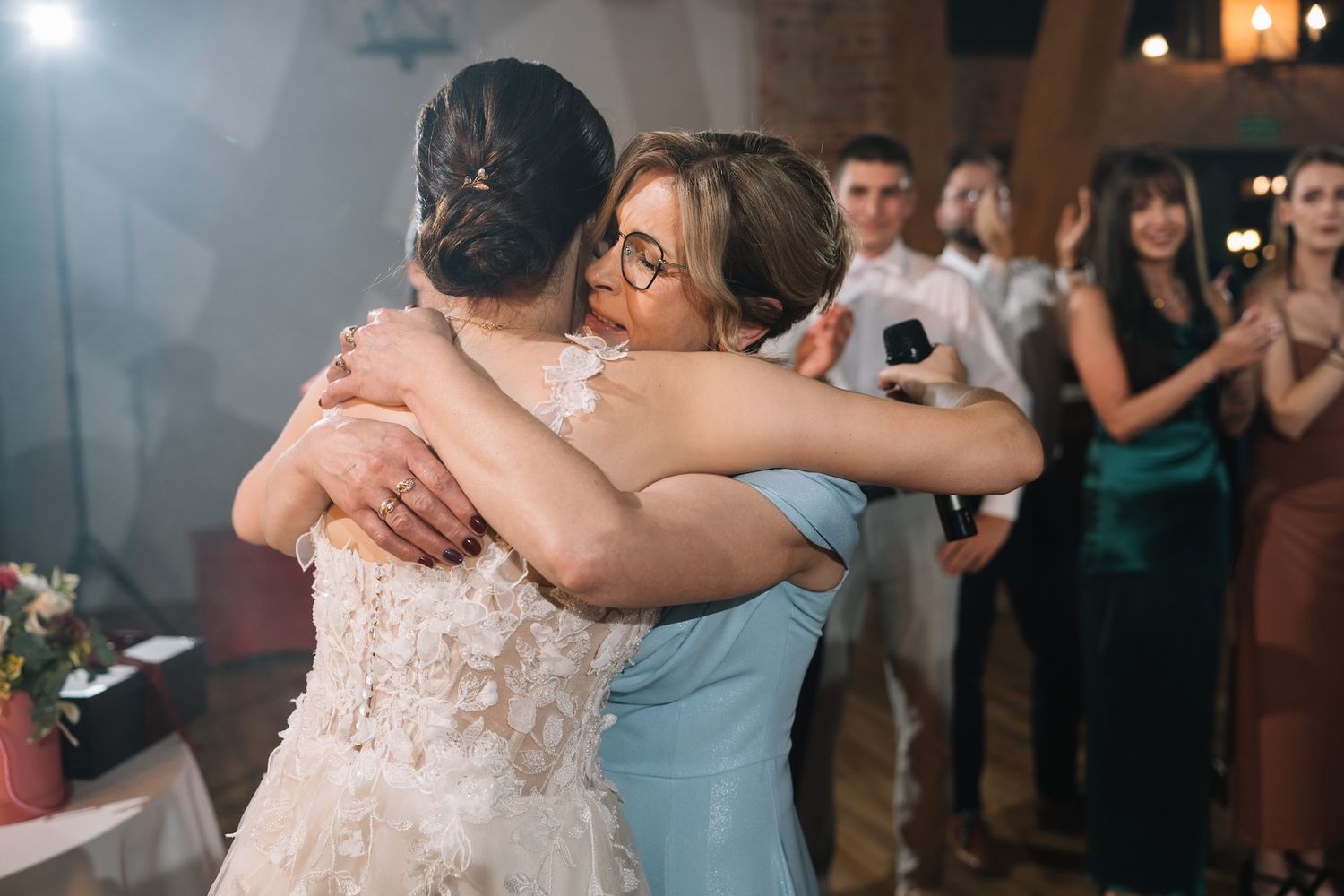 Bride in lace wedding dress shares emotional hug with woman in blue gown at wedding reception.