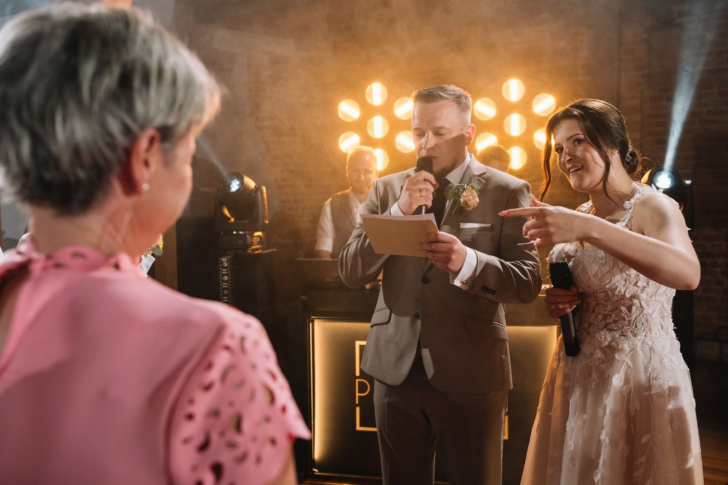 Groom in tan suit and bride clink glasses during wedding toast at reception with bokeh lights.