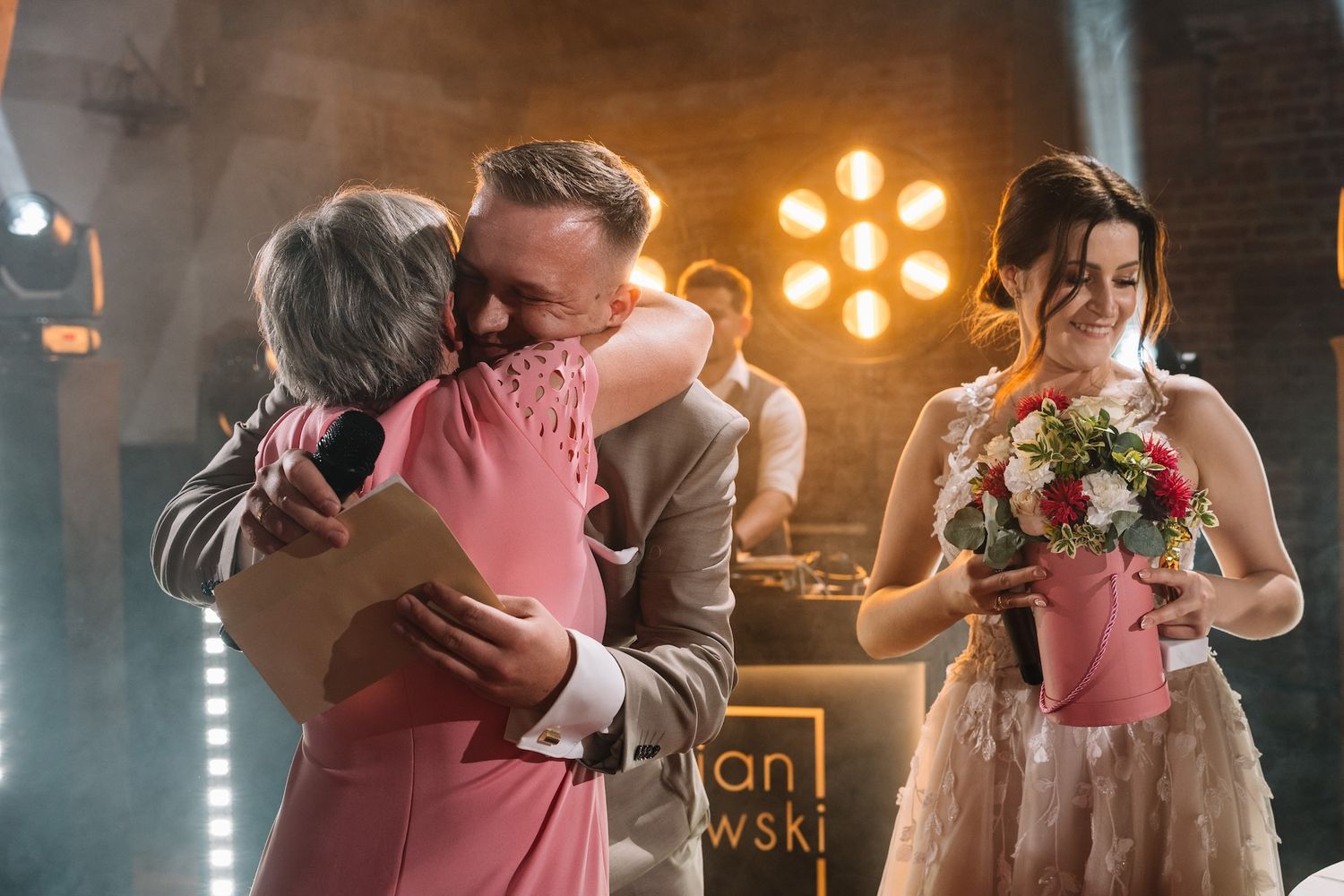 Groom embraces older woman in pink dress while bride holds bouquet at wedding reception venue.