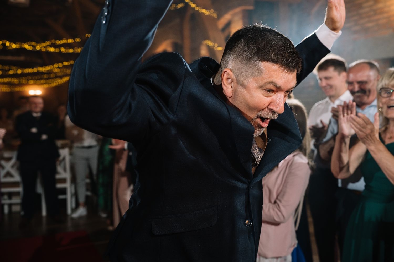 Man in dark suit energetically dances at lively wedding reception with guests clapping behind him.