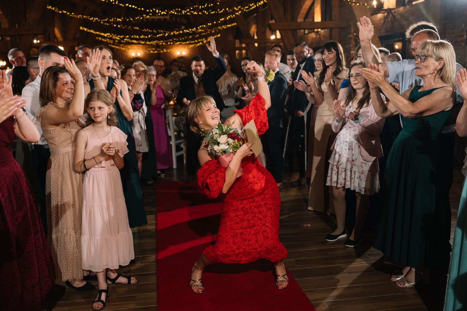 Woman in red dress catches bridal bouquet on red carpet at wedding reception surrounded by guests.