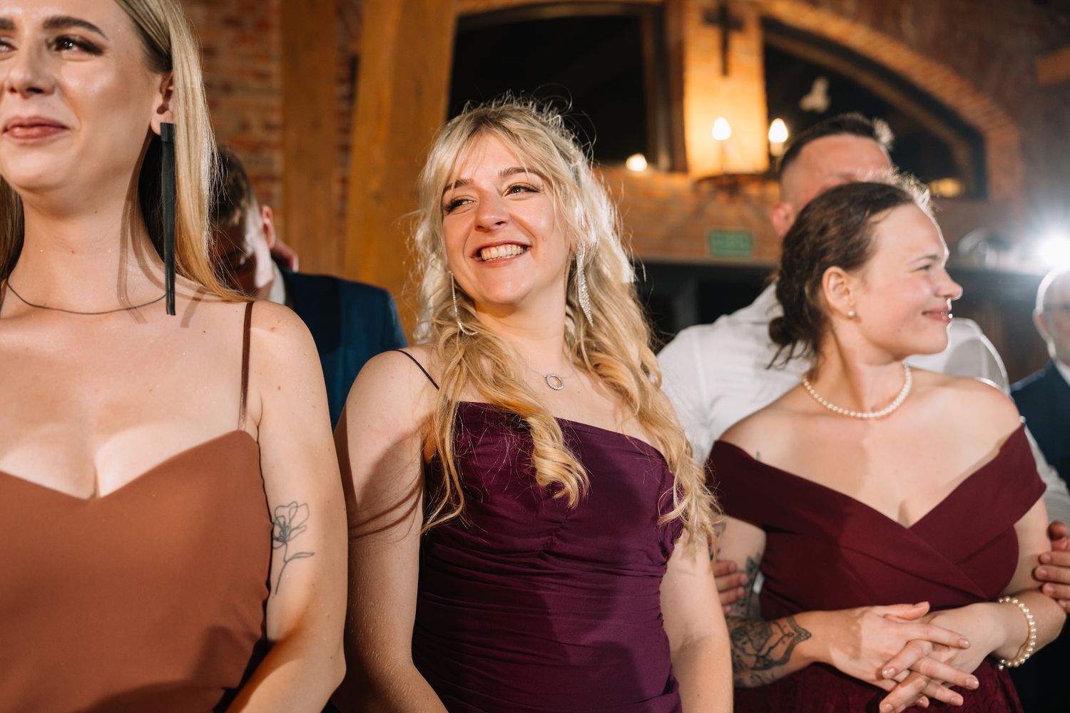 Three smiling women in elegant dresses enjoy wedding reception in rustic venue with brick walls.