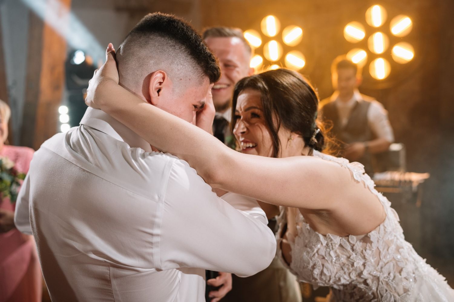 Bride in lace gown laughs joyfully while embracing groom at emotional wedding reception moment.