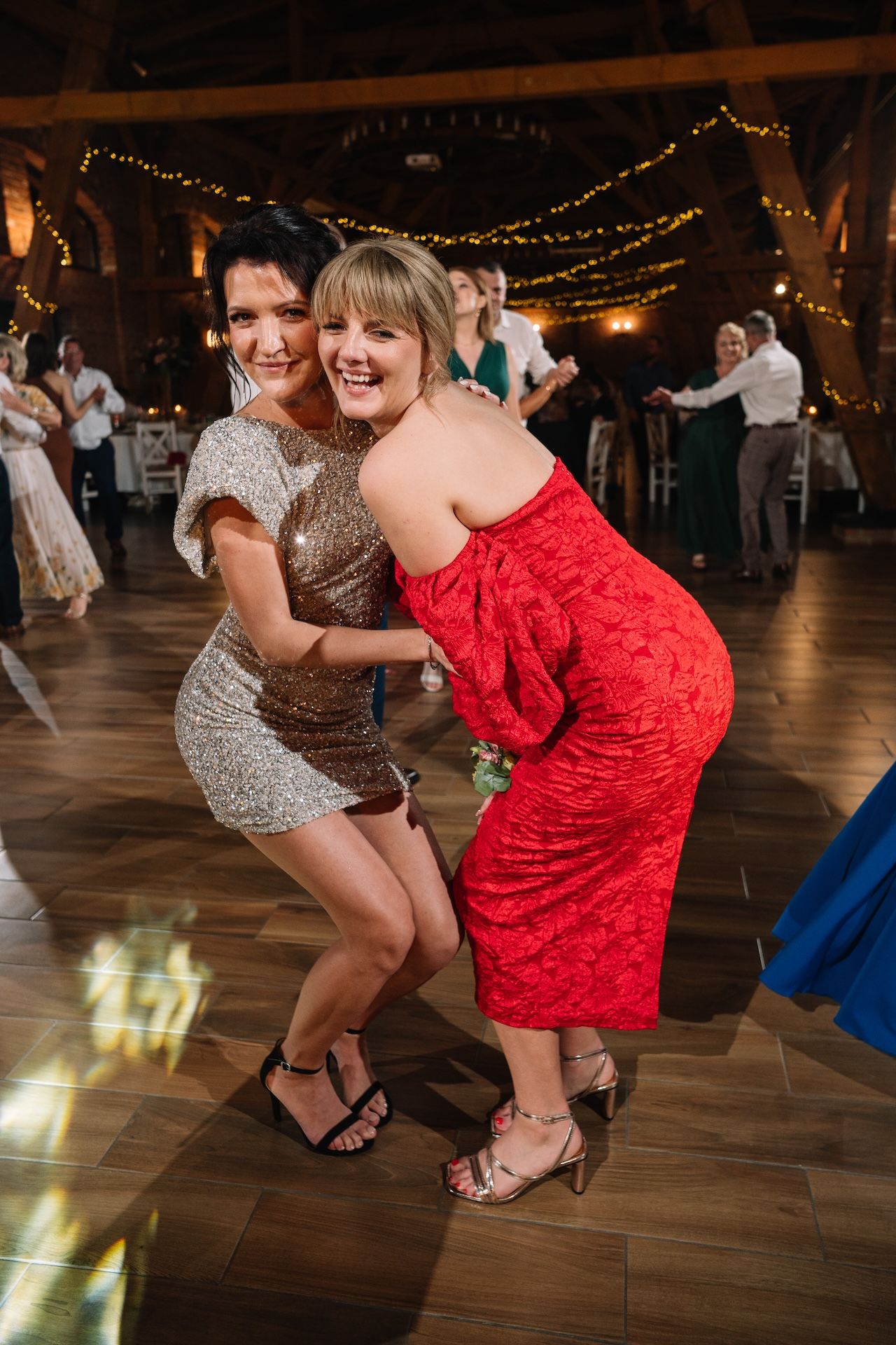 Two women in sequin and red dresses pose and dance together at lively wedding reception.