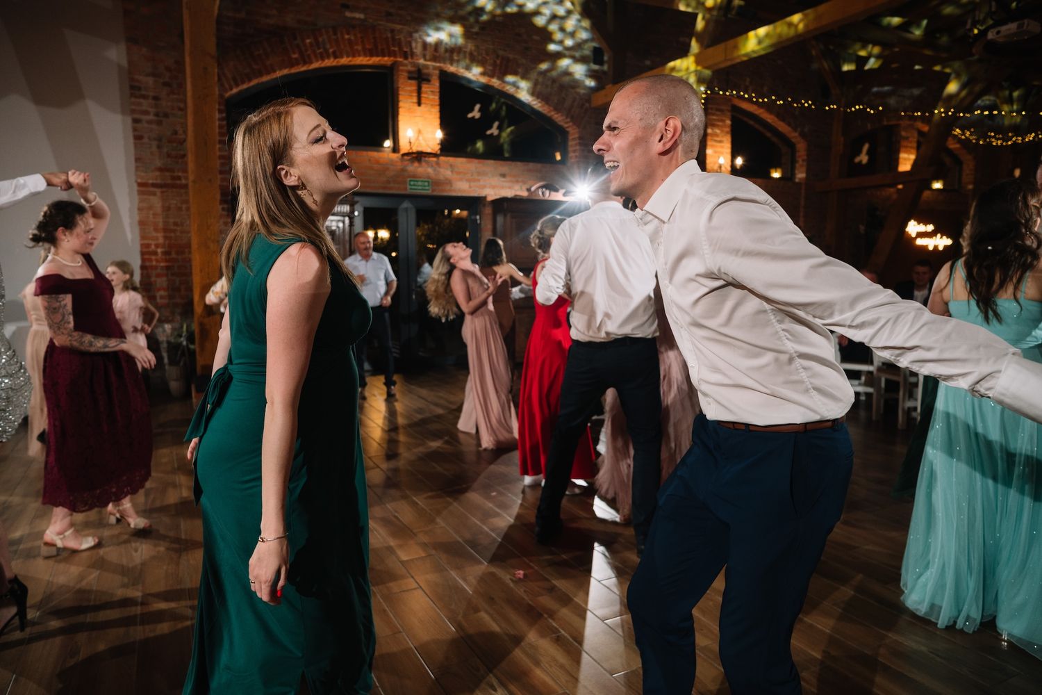 Couple in green dress and white shirt dance energetically at festive wedding reception celebration.