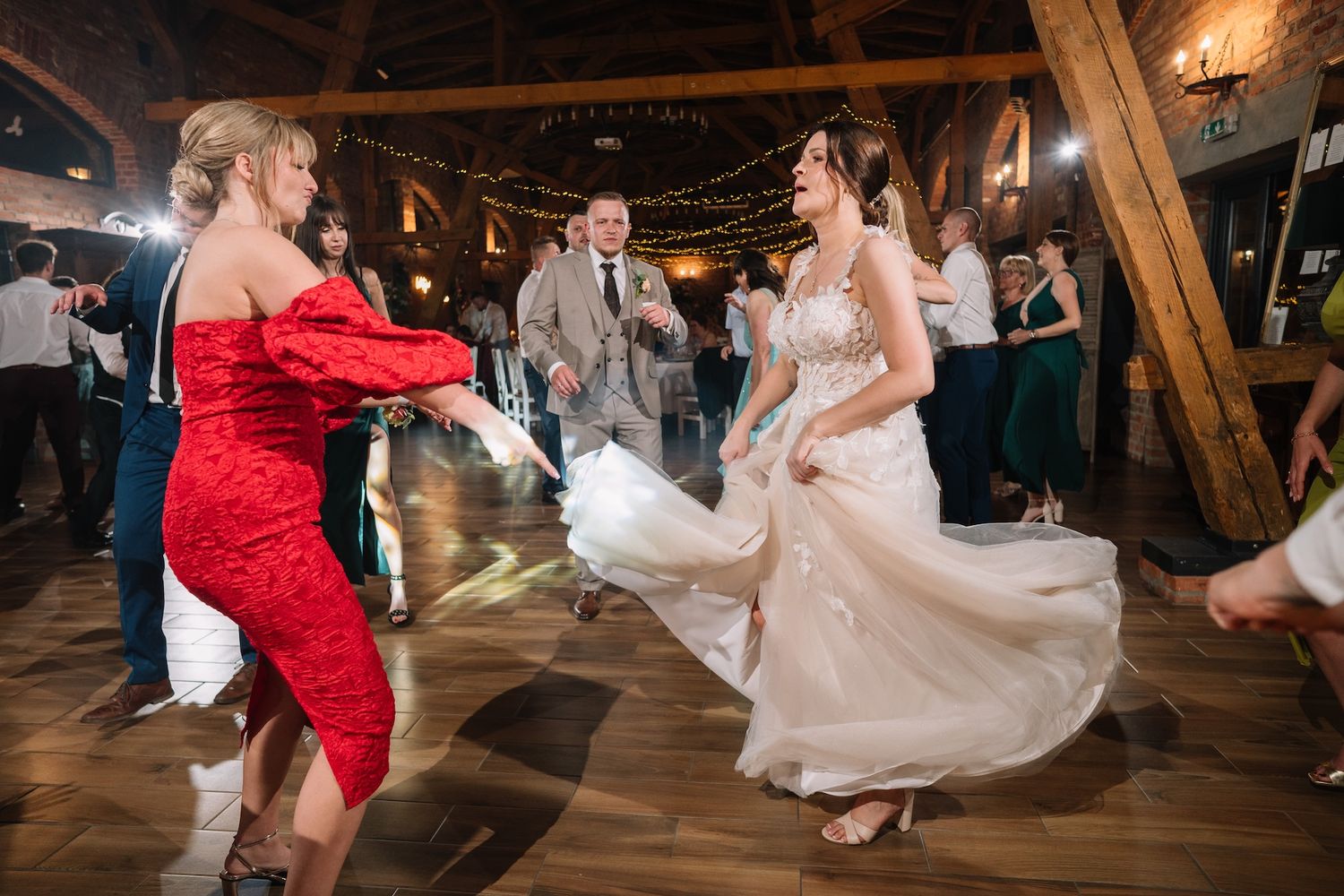 Bride in white gown twirling joyfully with guest in red dress on wedding reception dance floor.