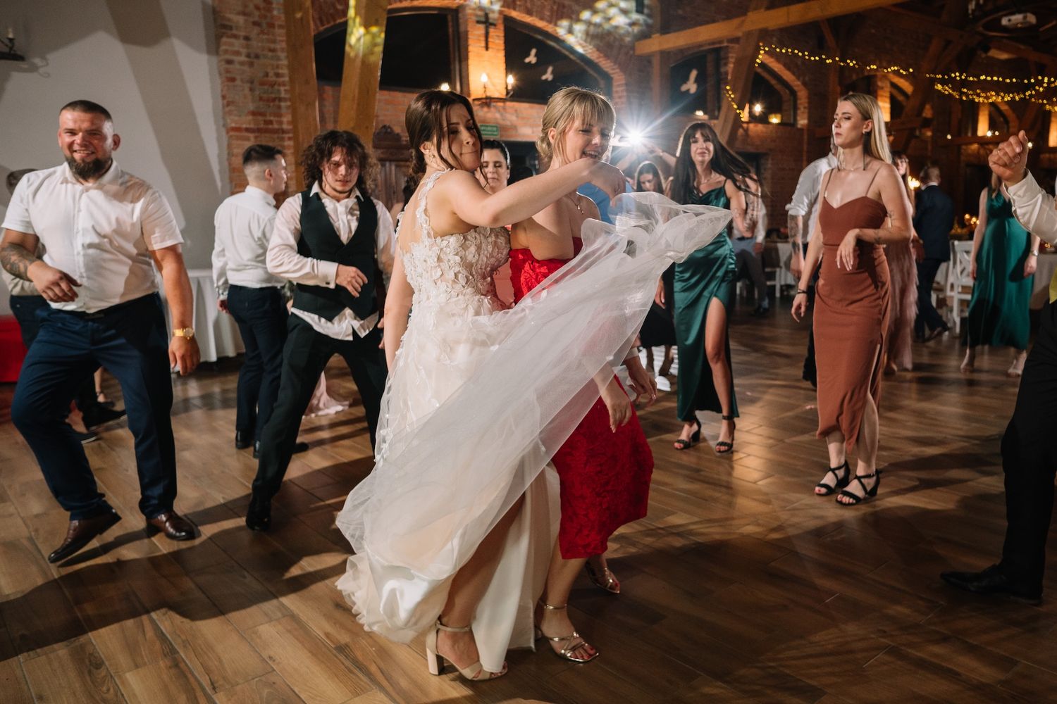 Bride dancing energetically with guests in elegant attire at a warmly lit rustic wedding reception.