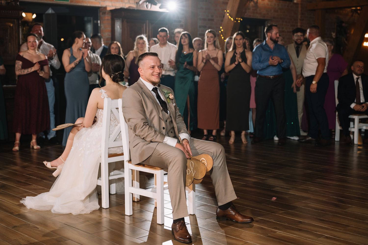 Bride and groom seated on white chairs smiling as wedding guests applaud during reception game.