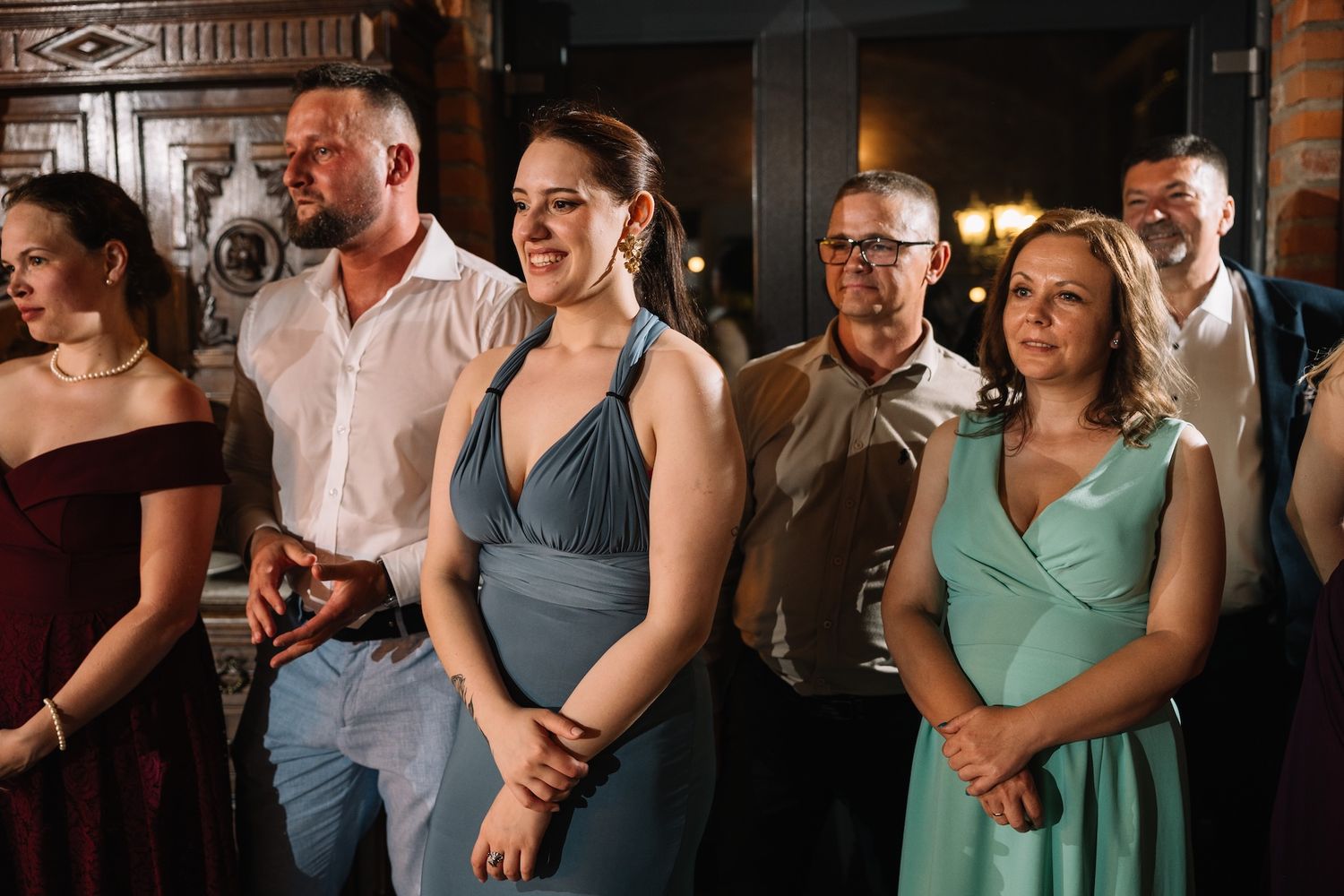 Smiling wedding guests in formal attire watching reception activities in a warmly lit venue.