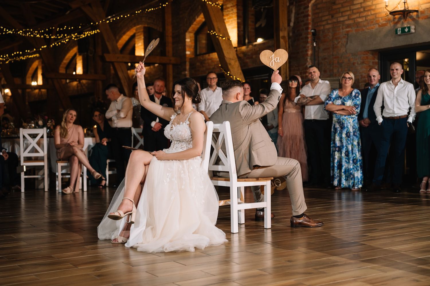 Bride and groom seated on chairs holding heart sign during fun wedding reception game.