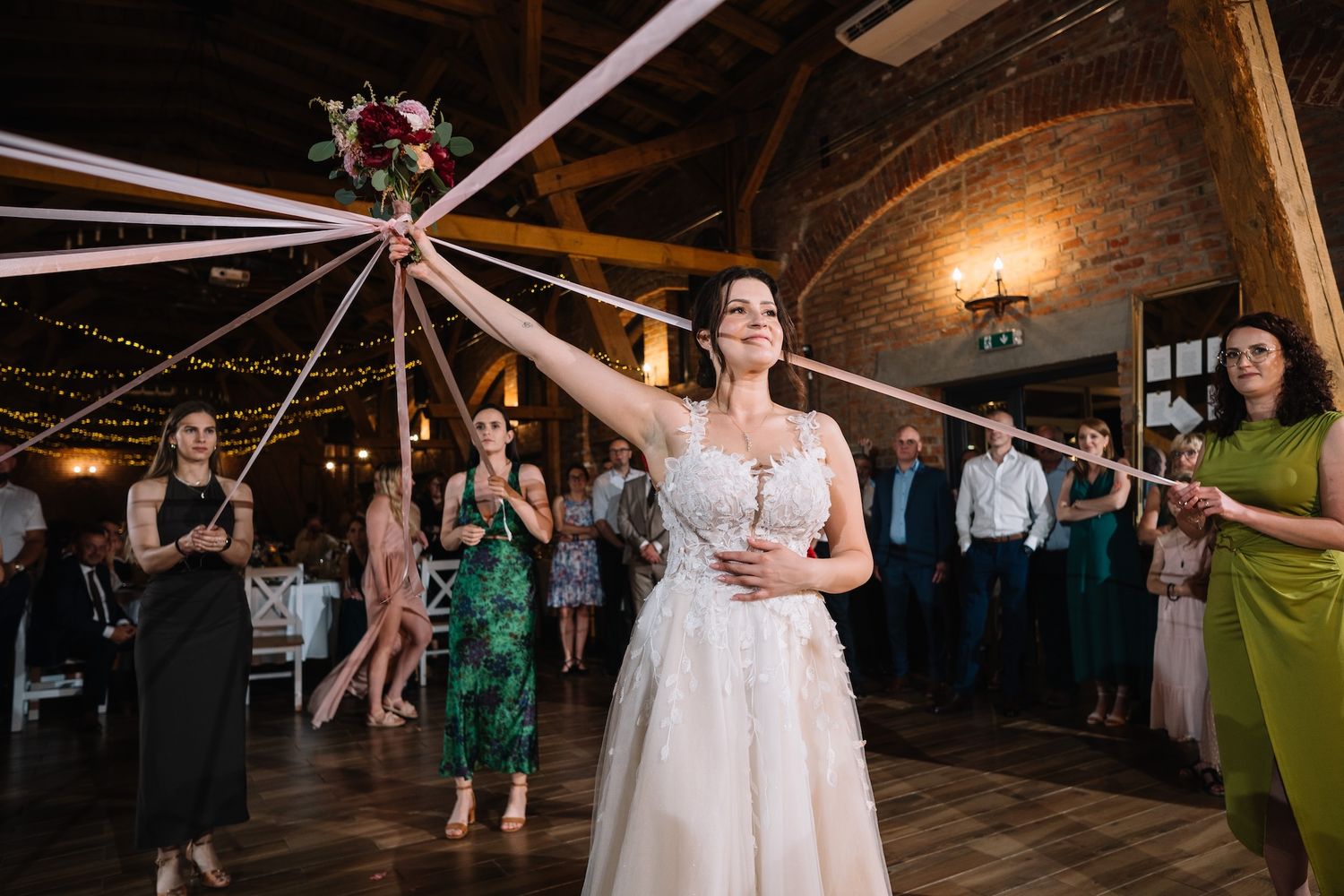 Bride in white gown holds bouquet with ribbons during wedding bouquet toss ceremony indoors.