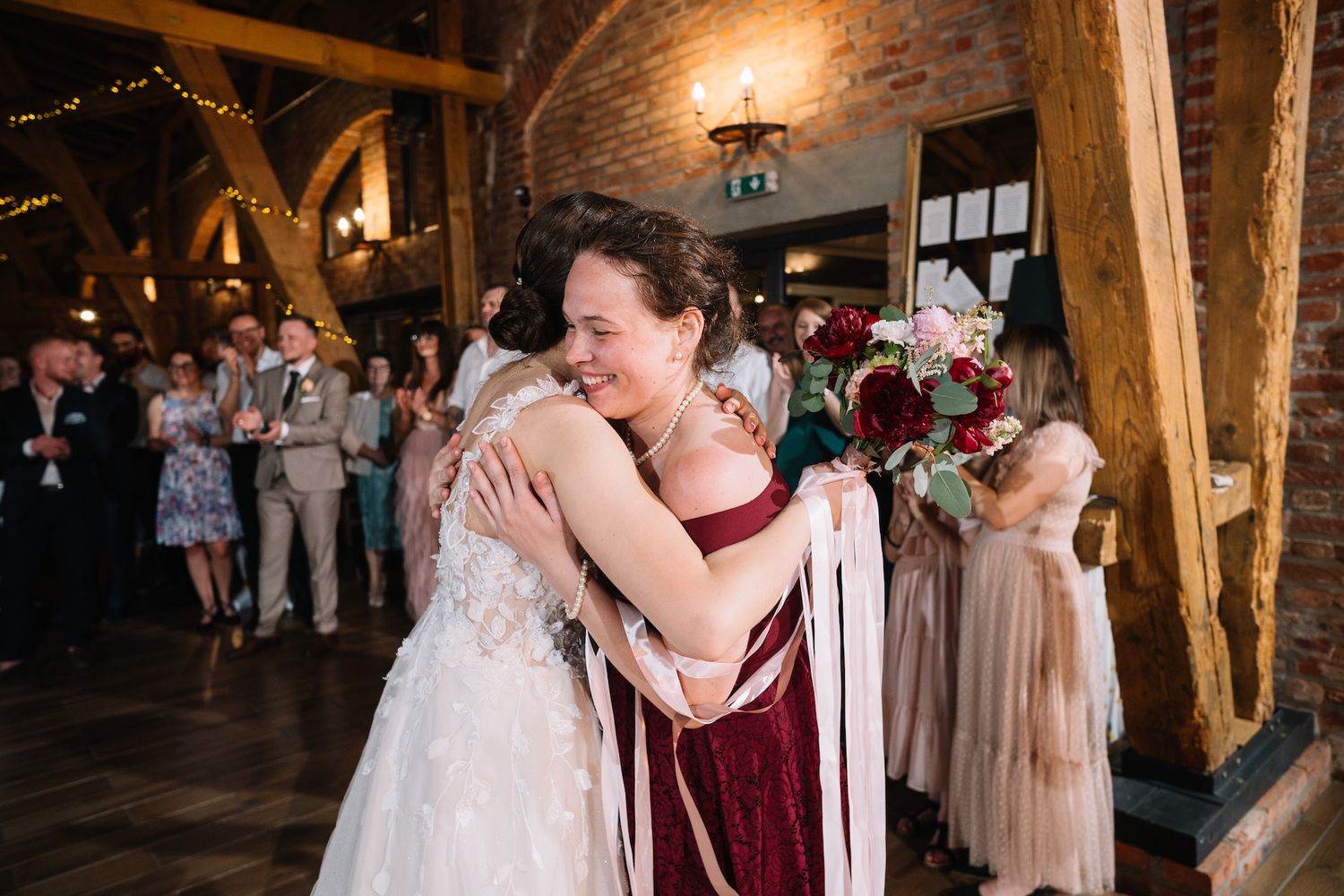 Bride in lace gown embraces bridesmaid in burgundy dress after bouquet toss at wedding reception.