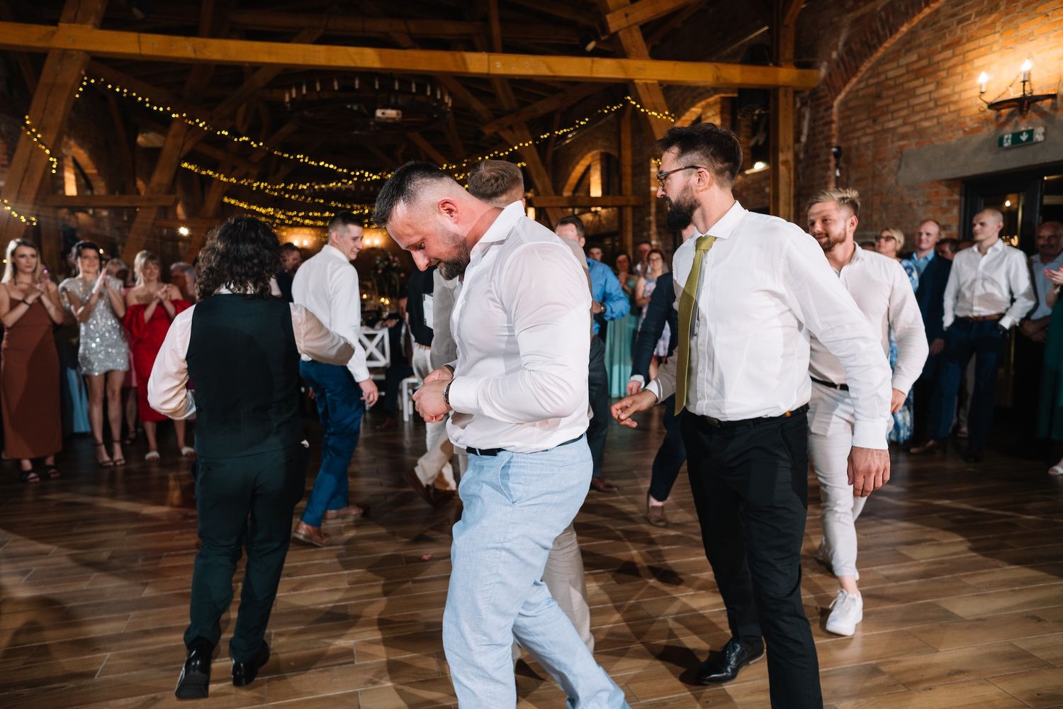 Groomsmen dancing energetically on wooden dance floor at rustic barn wedding reception.