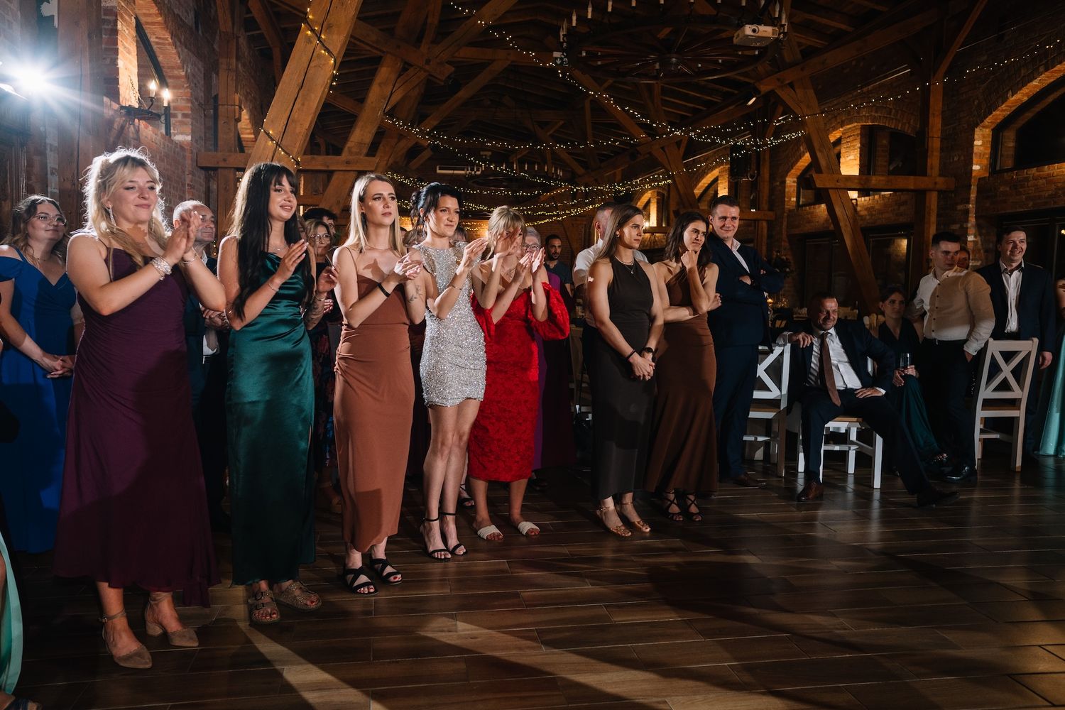 Elegantly dressed wedding guests in colorful gowns applaud on dance floor at rustic barn reception.