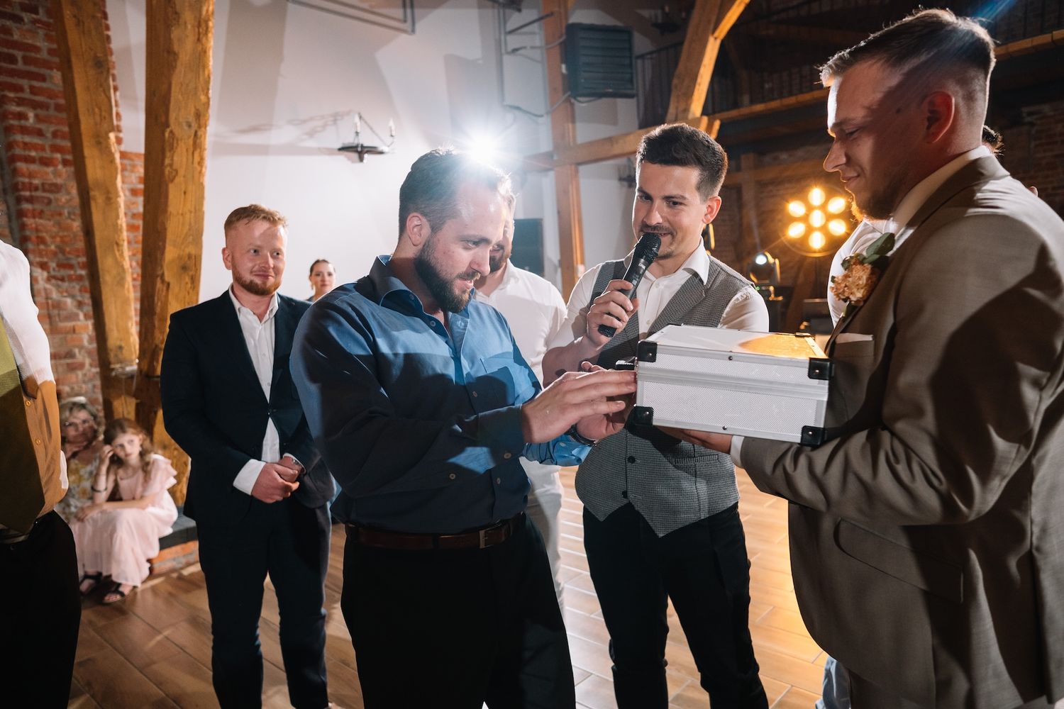 Men in formal attire examine item together under spotlight at rustic brick wedding venue.